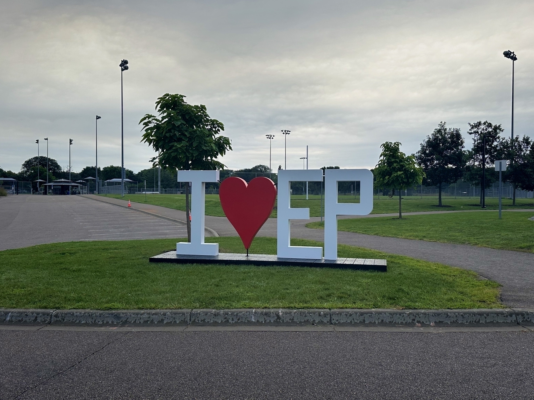 Large “I ♥ EP” sign resting on a grassy area, with trees and a paved road in the background, under a cloudy sky.