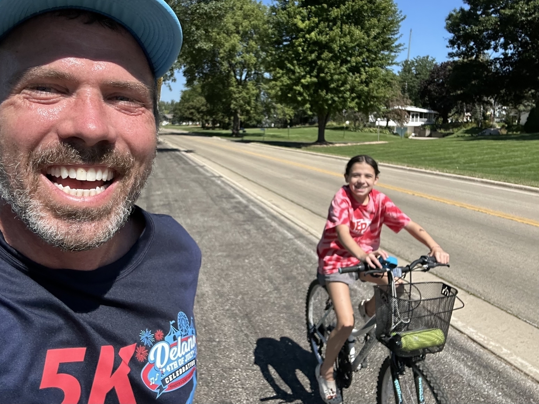 A man smiling at the camera while a girl rides a bicycle beside him on a paved road in a sunny park-like setting with trees and a house in the background.