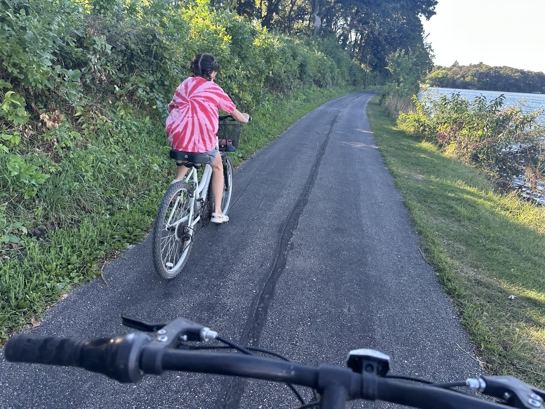 A person in a pink tie-dye shirt is cycling on a paved path next to a body of water and greenery, with another bike visible in the foreground.