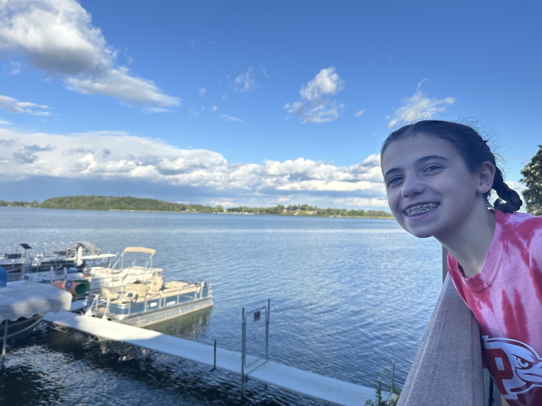 A smiling person with braces stands by a lake with boats docked nearby under a partly cloudy sky.