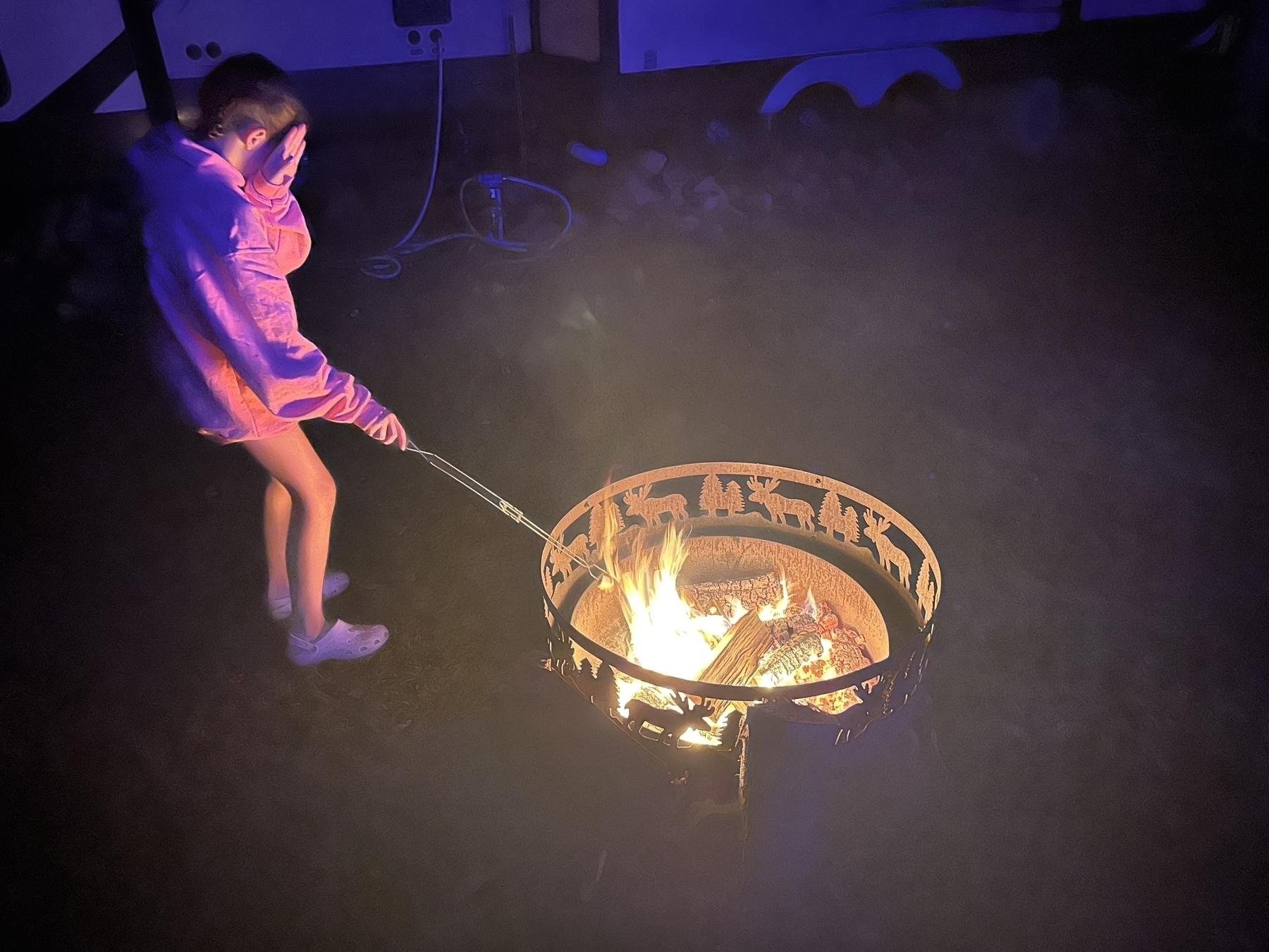 A person in a pink hoodie is roasting a marshmallow over a decorative fire pit at night.