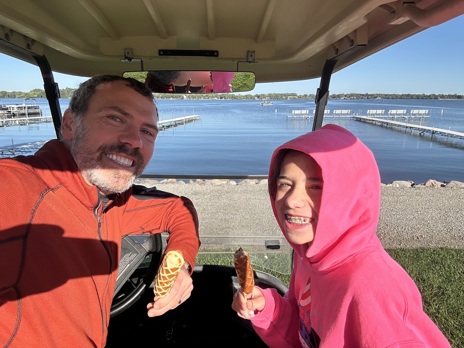 A man and a child are smiling while holding ice cream cones in a golf cart by a lake.