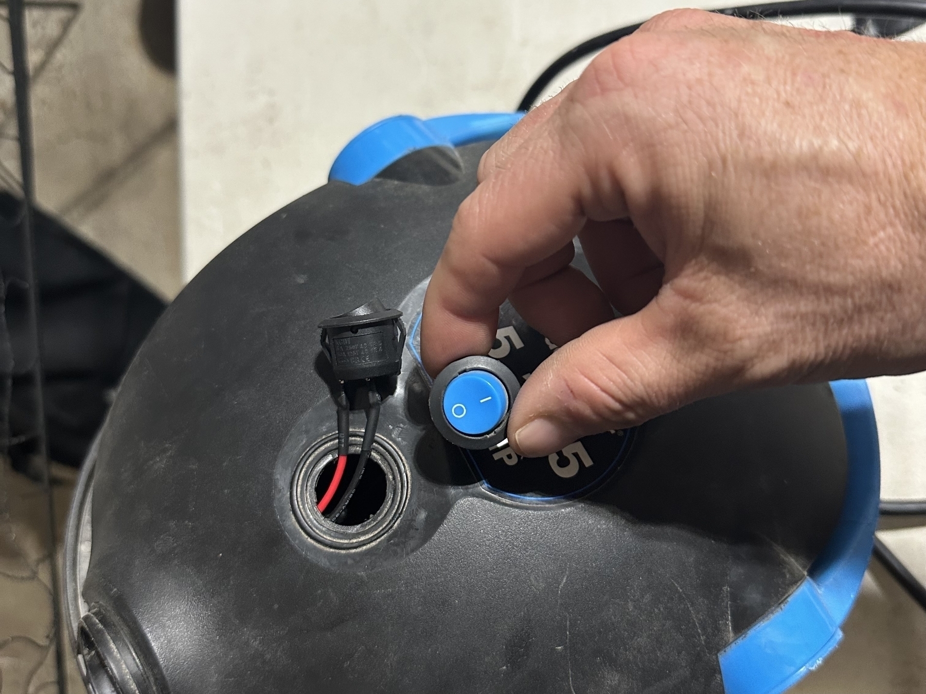 A hand installs a blue electrical switch into a hole on a black plastic surface next to an existing black plug; a concrete wall is in the background.