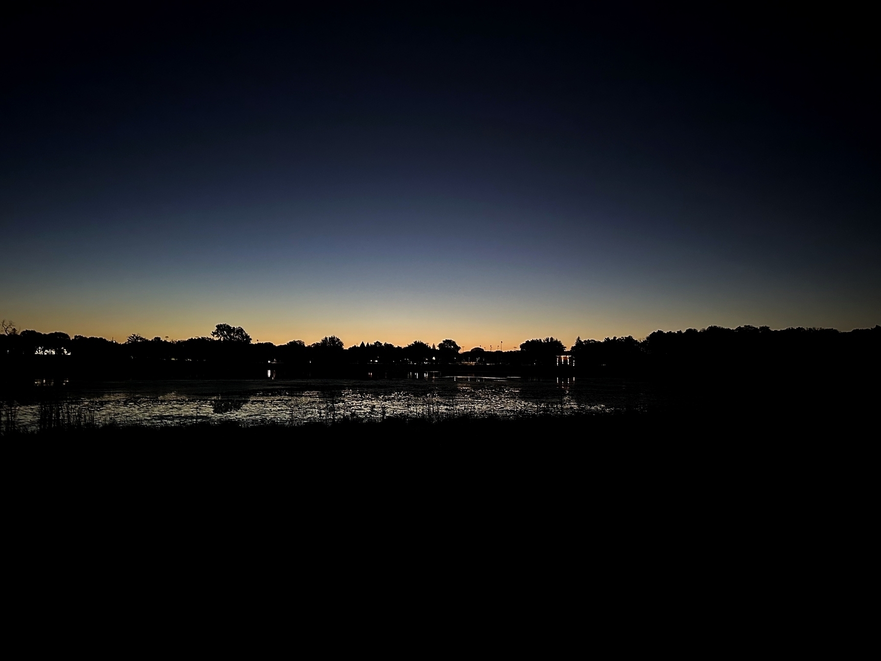 Silhouetted trees stand against a twilight sky, reflecting in a calm lake, creating a serene and tranquil evening scene.