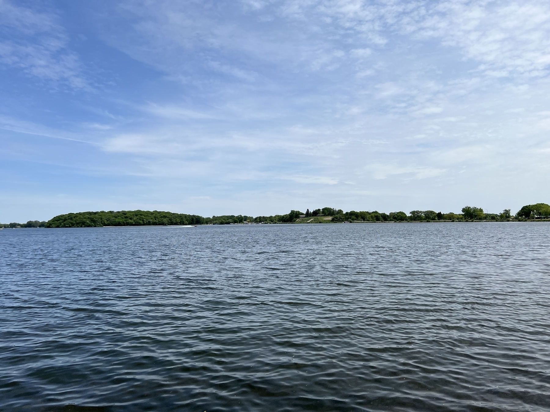 Calm water body resting under a partly cloudy sky with a forested hill on the distant horizon and scattered trees lining the shore.