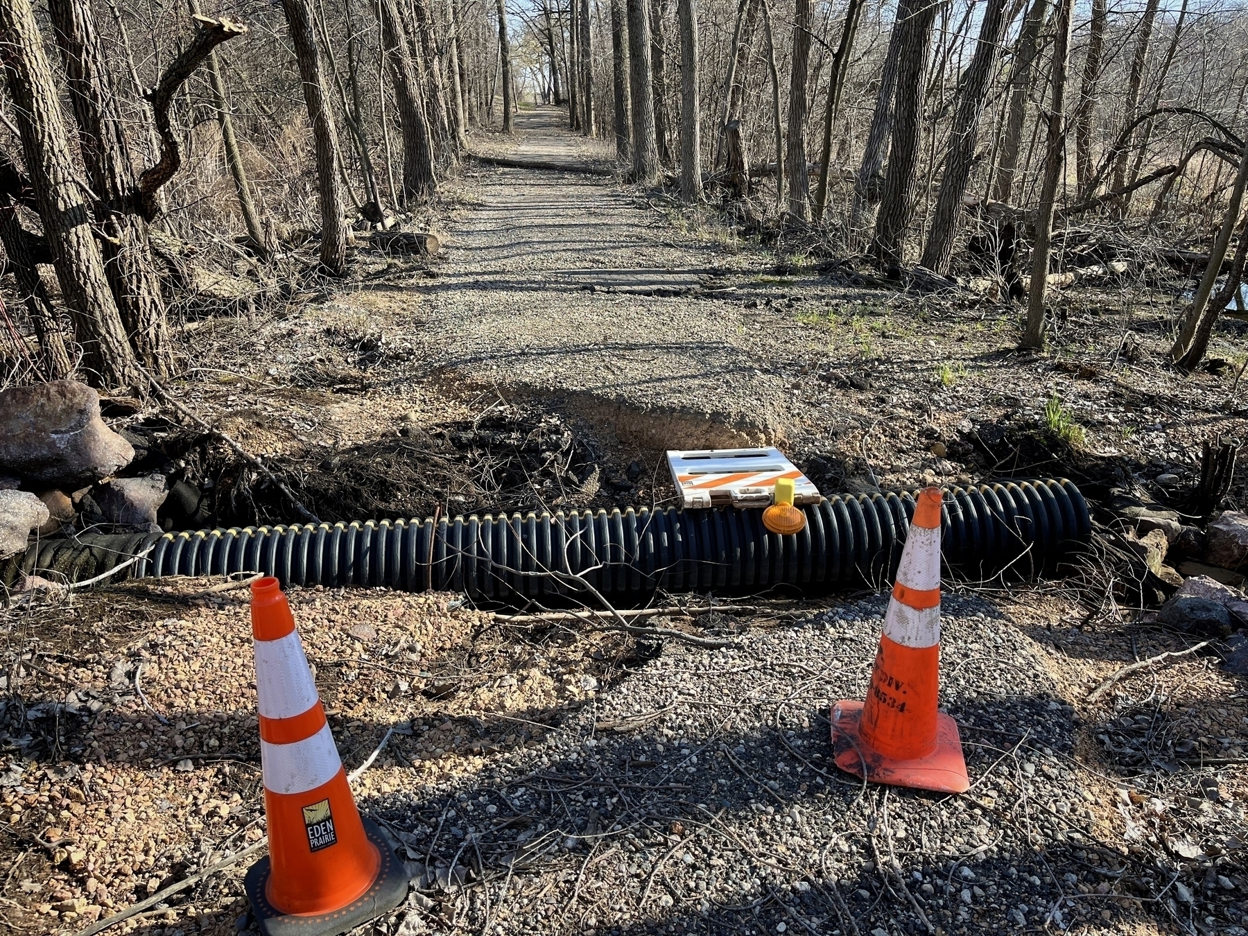 Traffic cones flank a corrugated pipe crossing a gravel trail in a wooded area, indicating ongoing maintenance or construction work.