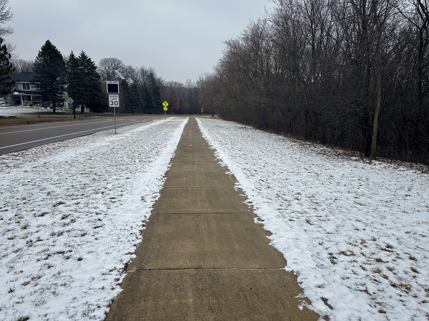 sidewalk flanked by grassy areas covered in snow, with a speed limit sign to the left and bare trees in the background