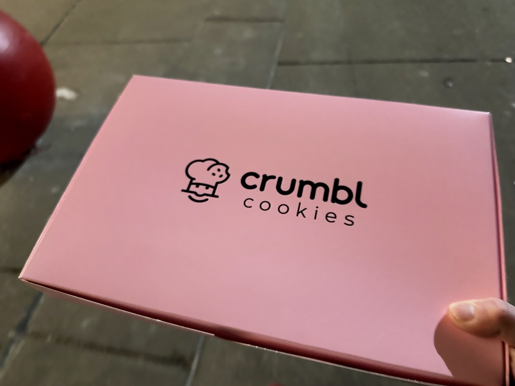 A hand holds a pink box with the logo “crumbl cookies” featuring a stylized cookie illustration, against a blurred street background