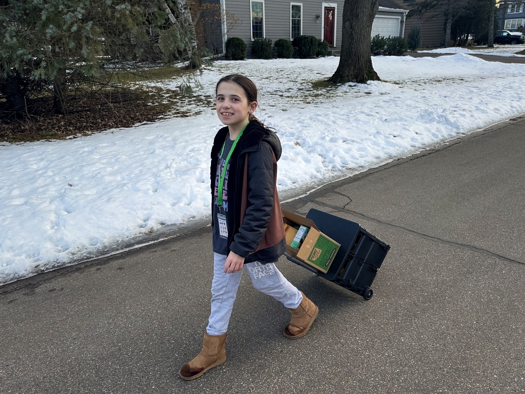 A young girl walks on a street, pulling a box on wheels, in a snowy suburban setting.
