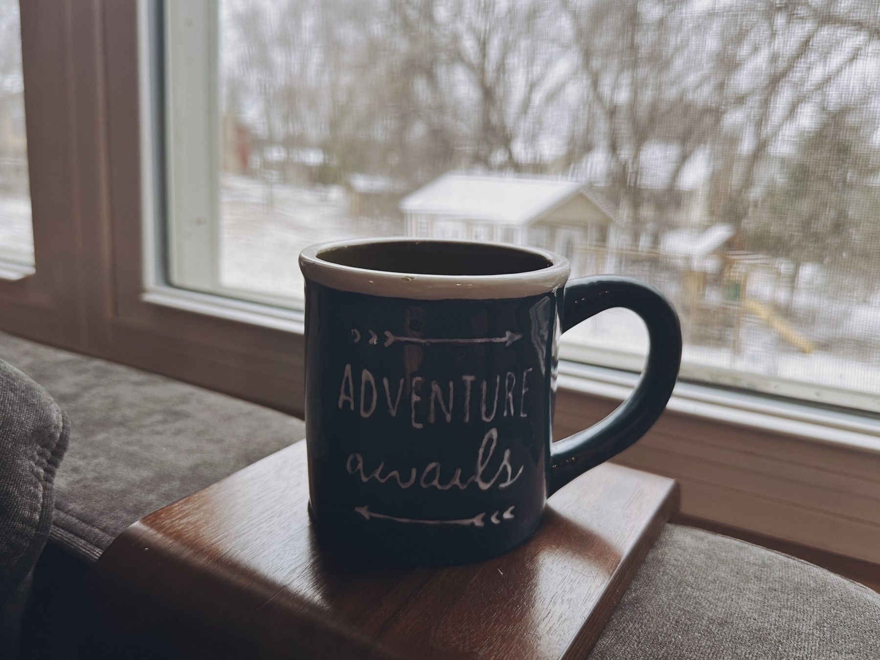 A mug with “ADVENTURE awaits” text is resting on a wooden surface, with a snowy residential view seen through a window in the background.