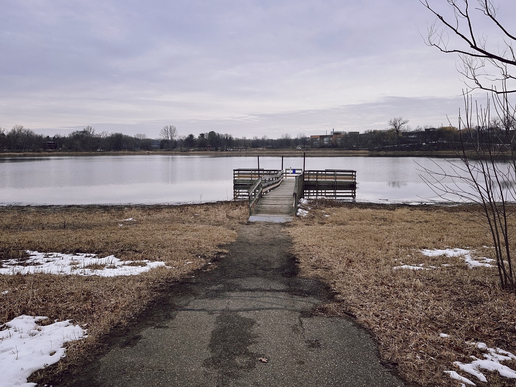 A weathered wooden pier extends into a calm lake with dry, brown grass and patches of snow along the shore, under a cloudy sky.