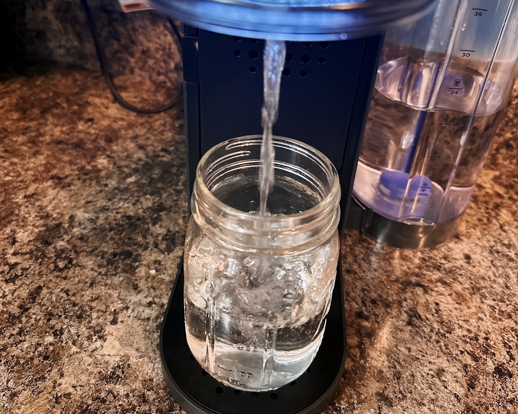A water filter pitcher is dispensing water into a glass jar on a kitchen countertop.