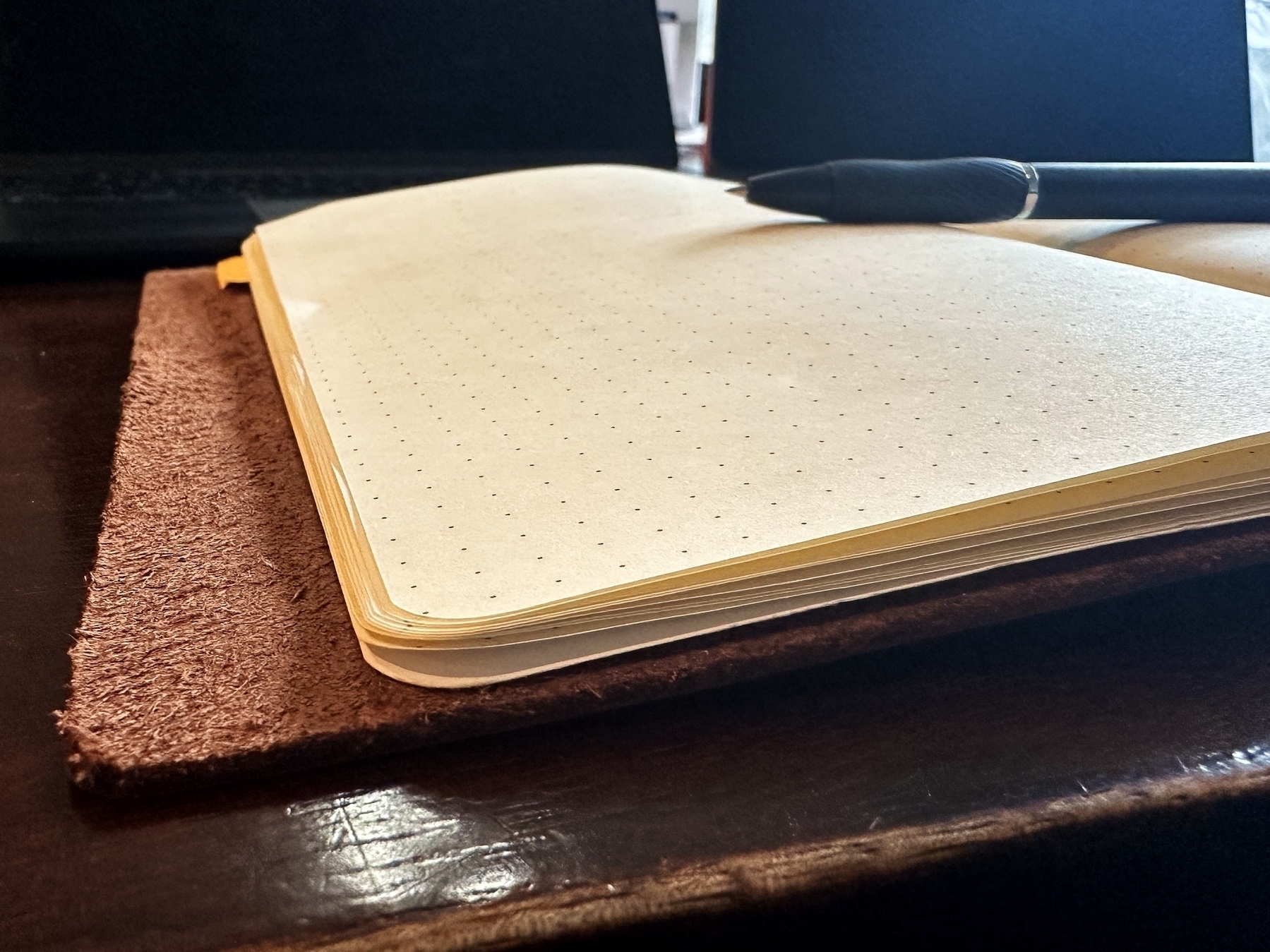 An open dot-grid notebook and pen rest on a leather surface next to a laptop, suggesting a work or study setting.