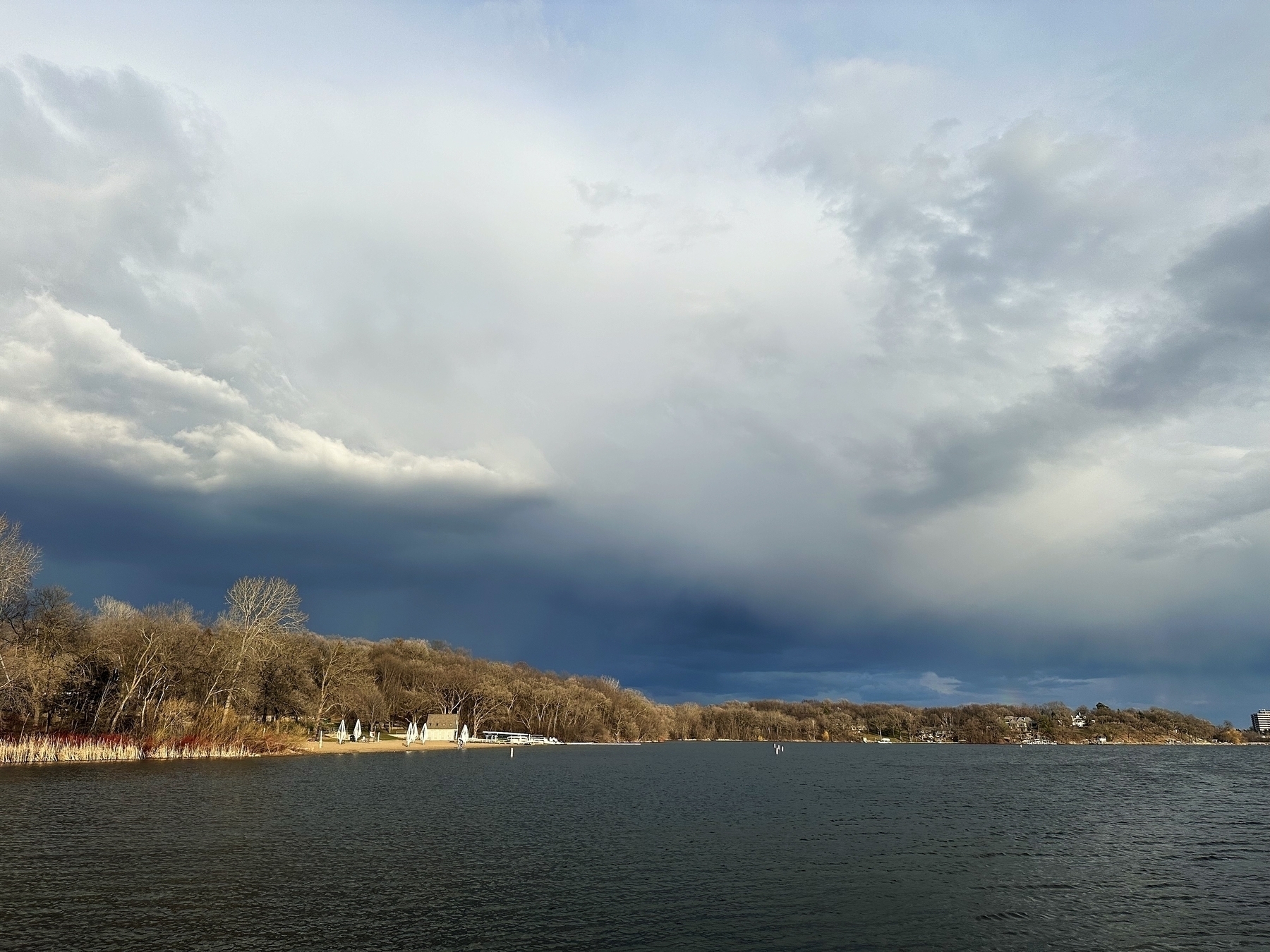 Overcast sky looms above a calm lake with bare trees and distant buildings lining the shore, suggesting an impending storm in a serene natural setting.