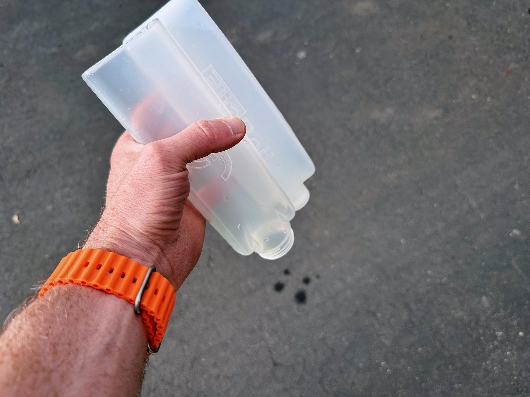 A hand holding an empty, translucent plastic bottle over a paved surface. The wrist is adorned with an orange watch strap.