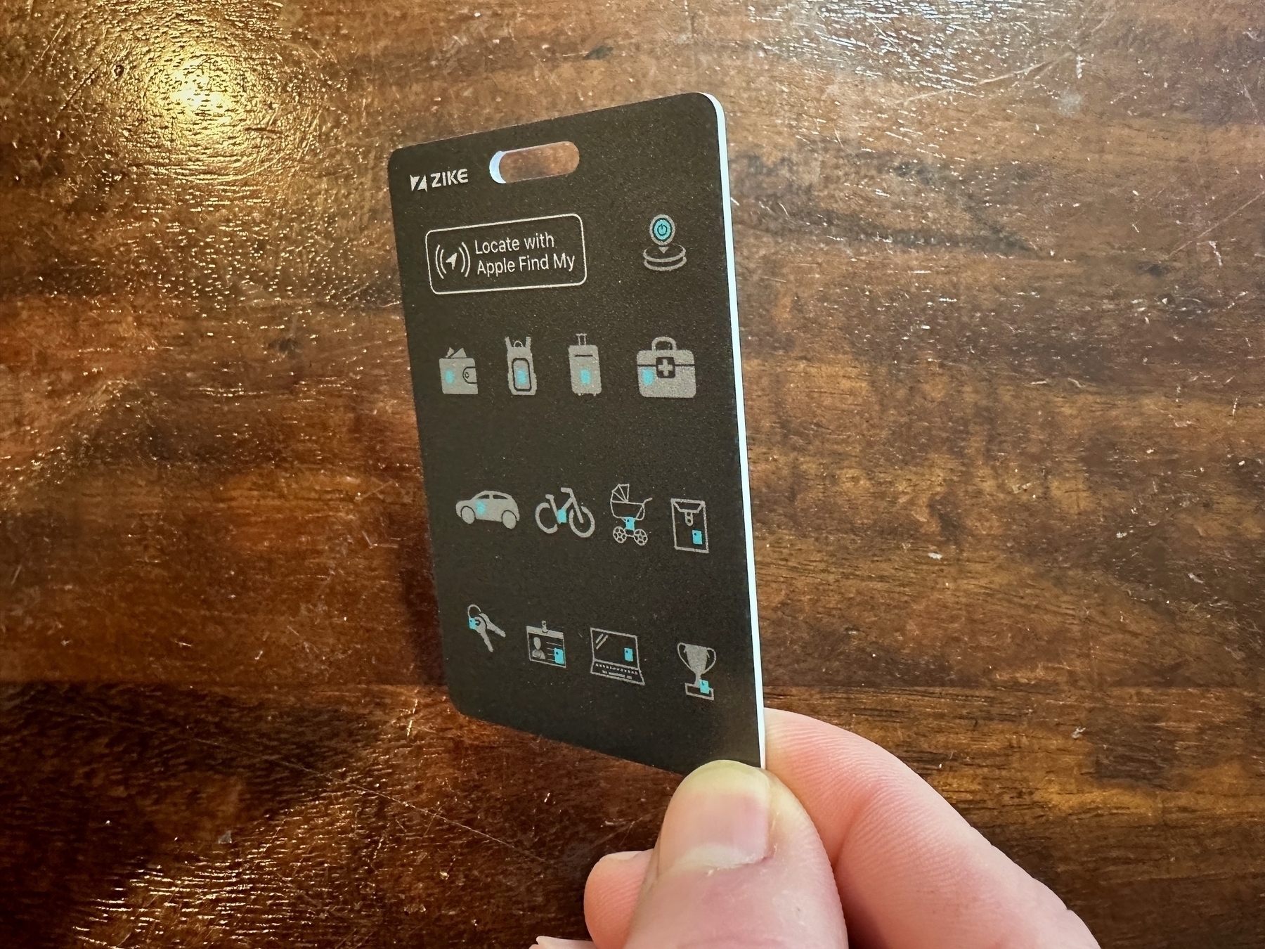 A hand holds a black card with icons and text that reads “ZIKE, Locate with Apple Find My” against a wood-grain background.