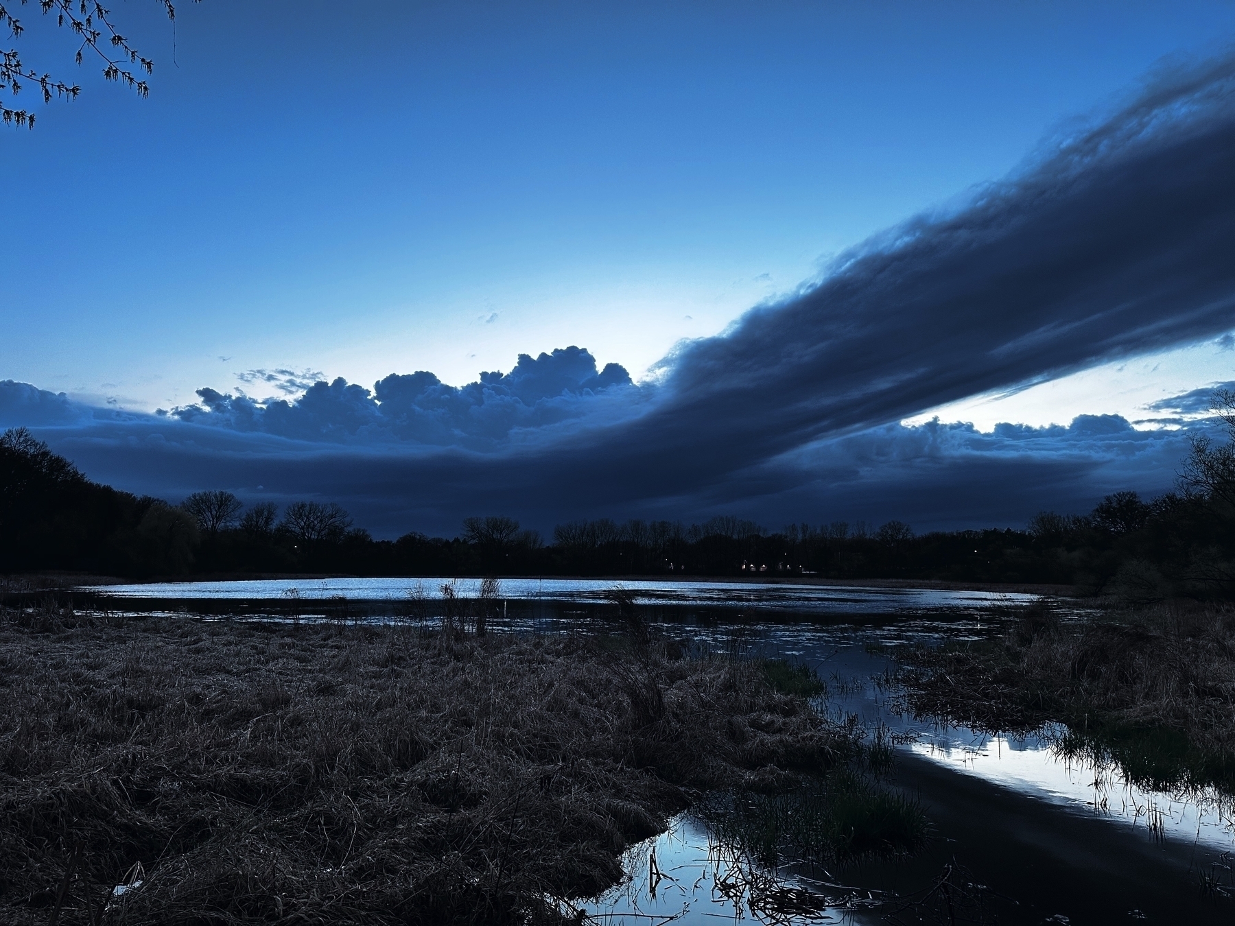 Dark clouds loom over a tranquil water body at dusk, reflecting the waning light, amidst a landscape of contrastingly tranquil fields and trees.