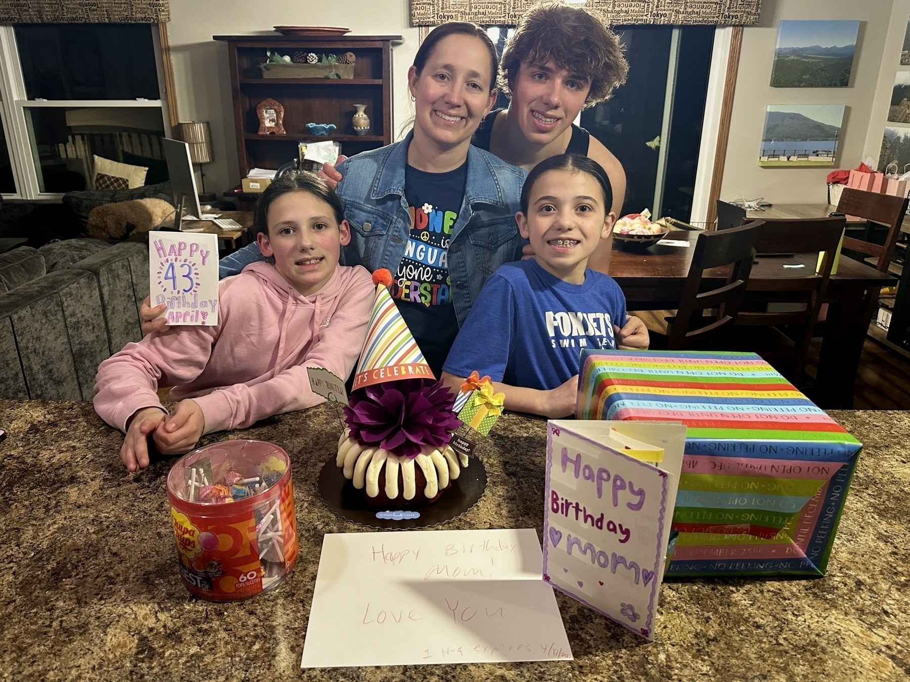 A family celebrates a birthday at home, surrounding a cake with presents and handmade cards reading “Happy 43rd Birthday April” and “Happy Birthday Mommy… Love You.”
