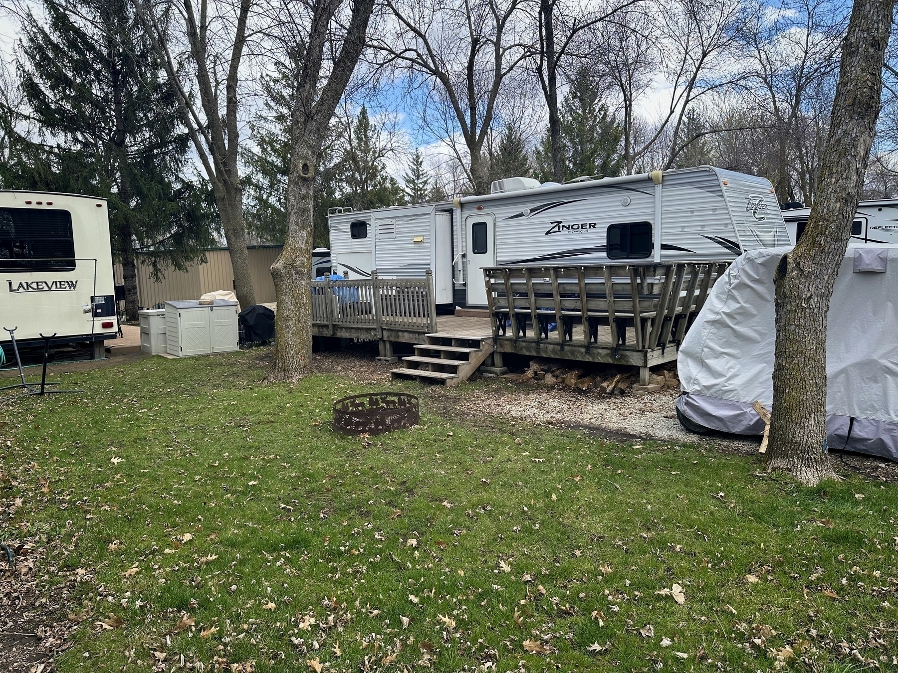 A stationary trailer with an attached wooden deck is set among leaf-scattered grass, flanked by trees and another trailer, indicating a campsite or RV park.