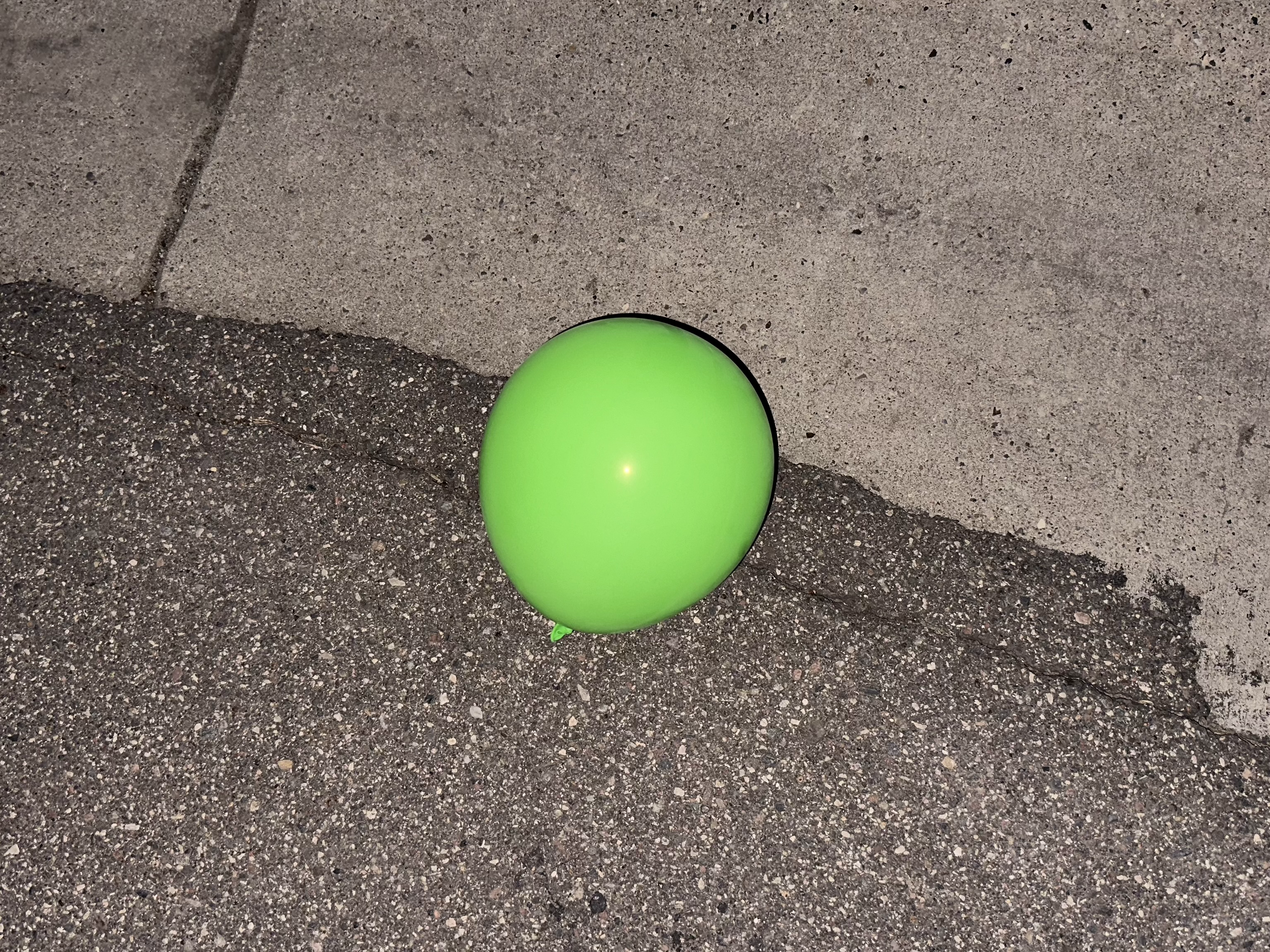 A green balloon rests on asphalt near a concrete curb under ambient light at night.