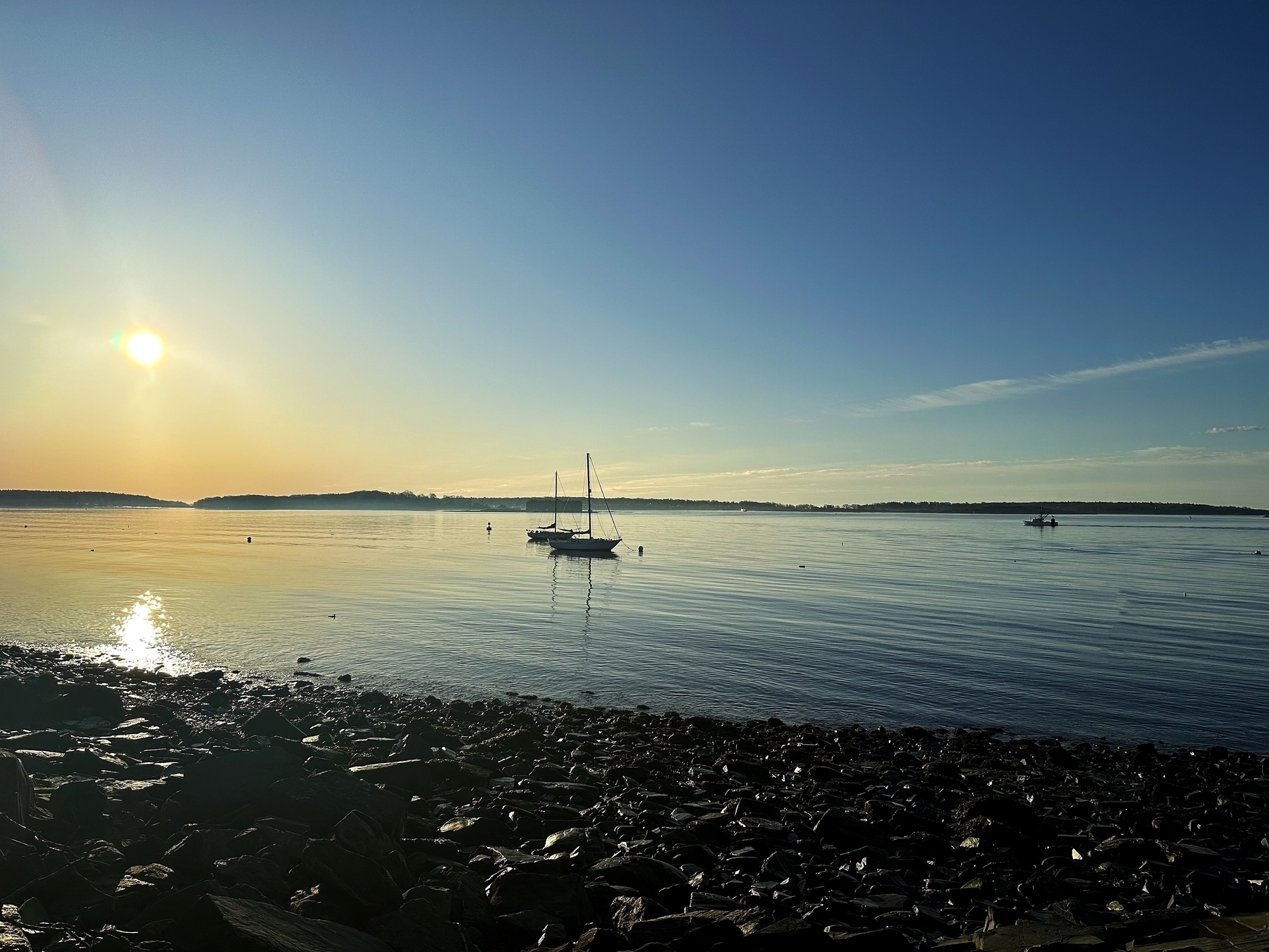 Sailboats rest on calm water at sunrise/sunset, with sun casting reflections; rocky shore in the foreground, clear skies above. No text is present.