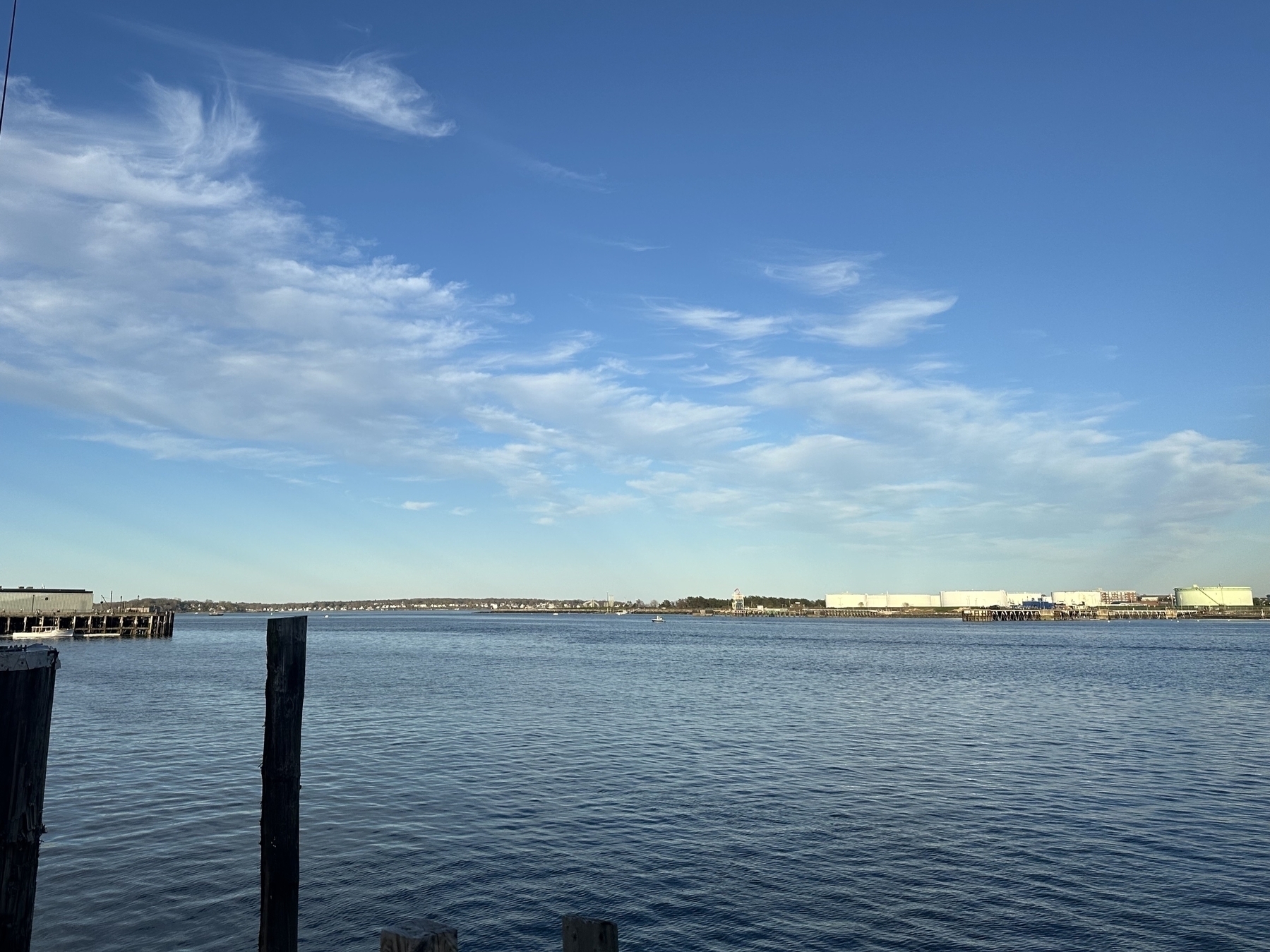 A serene waterscape under a blue sky with wispy clouds, bordered by industrial structures and a pier, suggesting a harbor or coastal area.