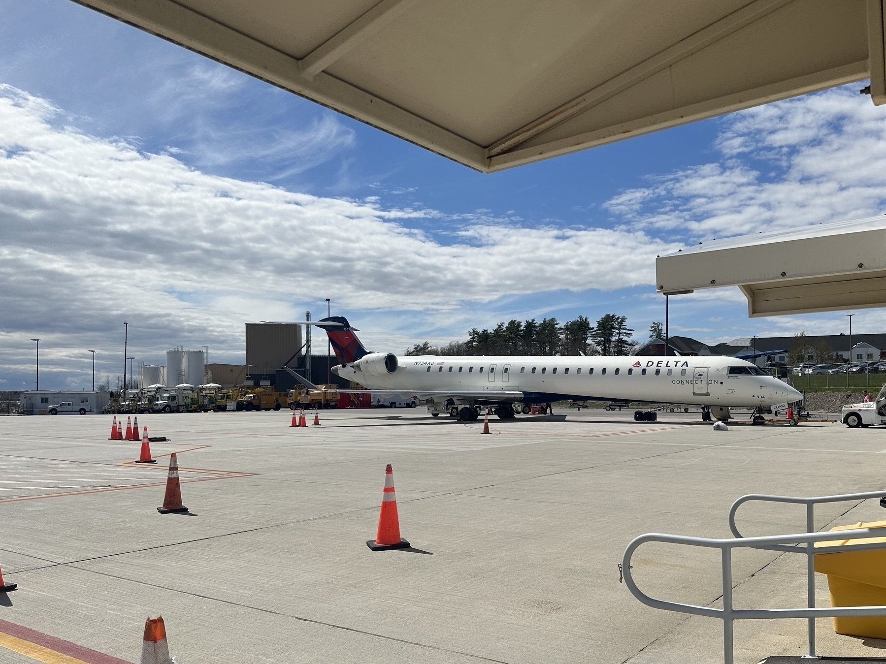 A Delta Connection aircraft is parked on the airport tarmac, with ground support equipment and vehicles nearby, under a partly cloudy sky.