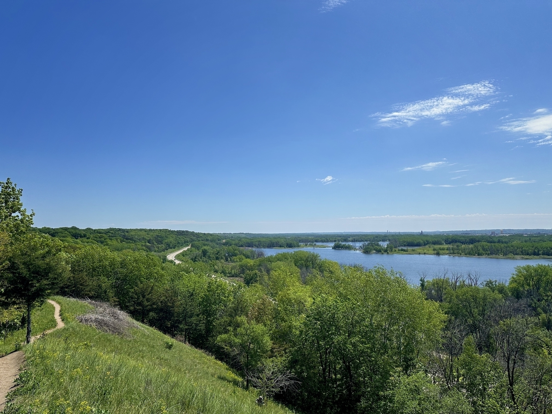 A path winds through a lush green hillside surrounded by dense trees with a river positioned below and the vast blue sky above.