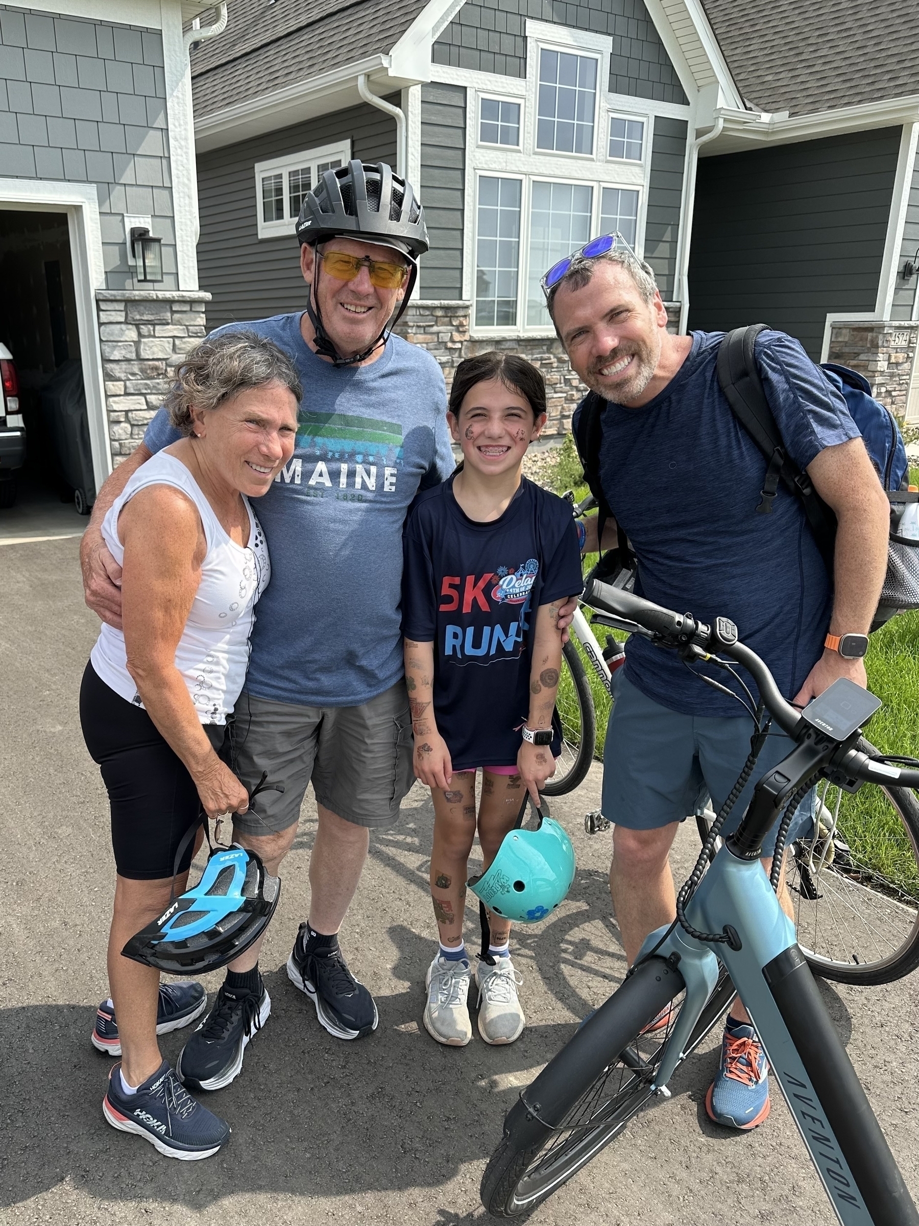 Four people smiling, standing in front of a house: a man in a helmet and “Maine” shirt, a woman holding a helmet, a child holding a helmet, and a man with a bike. “5K RUN” on the child’s shirt.