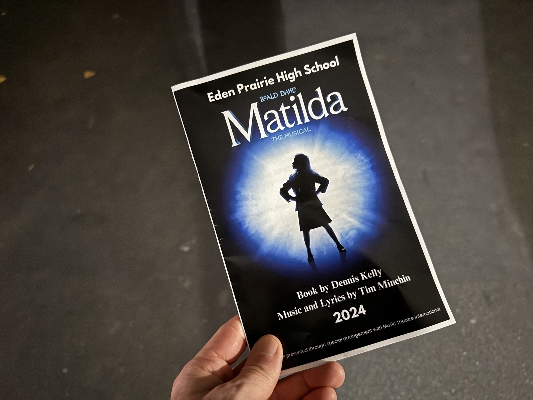 A person holds a program for “Matilda the Musical” presented by Eden Prairie High School. The cover features a silhouette of a girl standing confidently against a blue glowing background. The program lists the book by Dennis Kelly and music and lyrics by Tim Minchin, dated 2024.