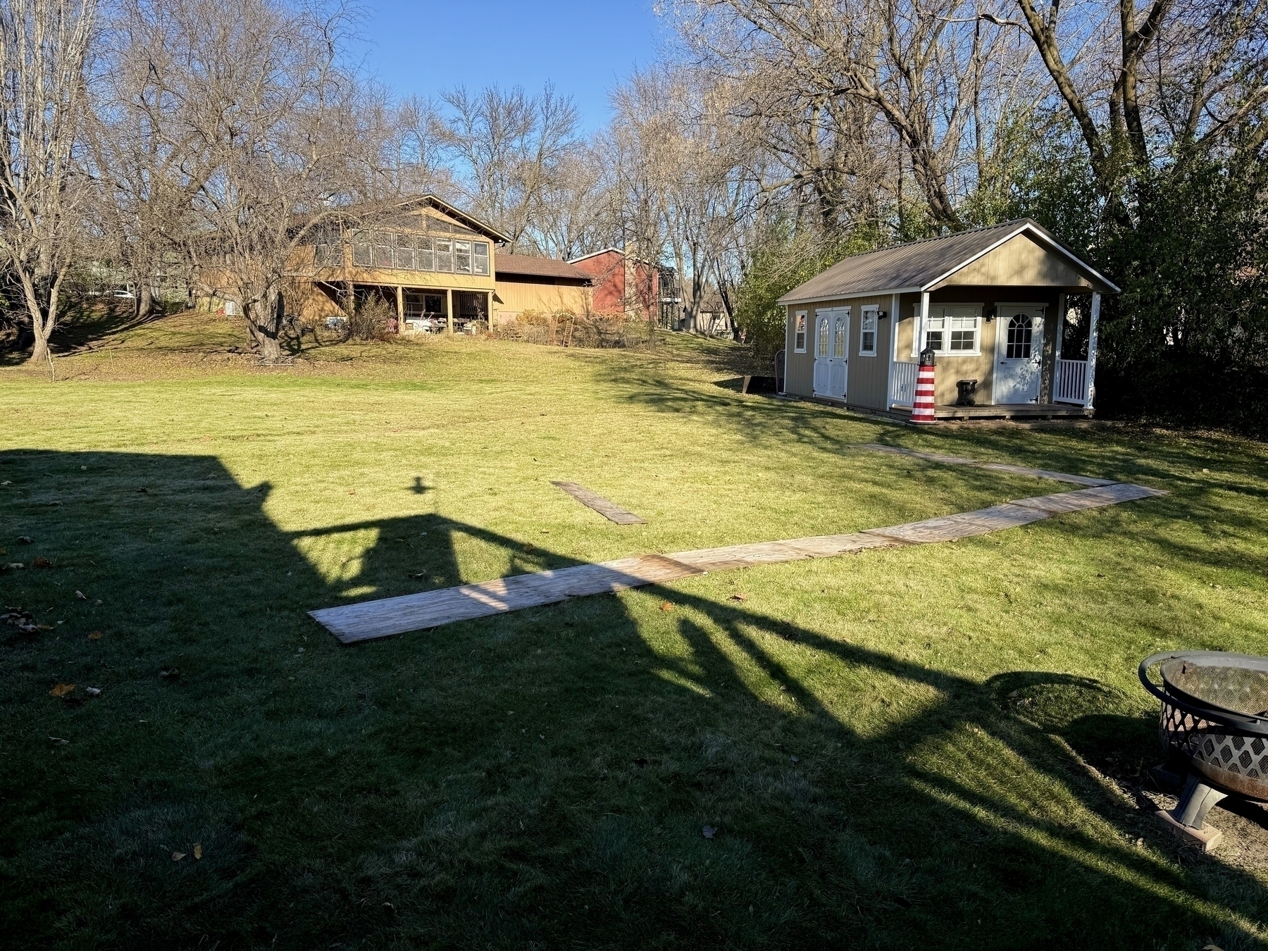 A grassy backyard area with a small shed on the right and a larger house in the background. A curved wooden path leads through the lawn. Bare trees surround the yard, with clear blue skies overhead.