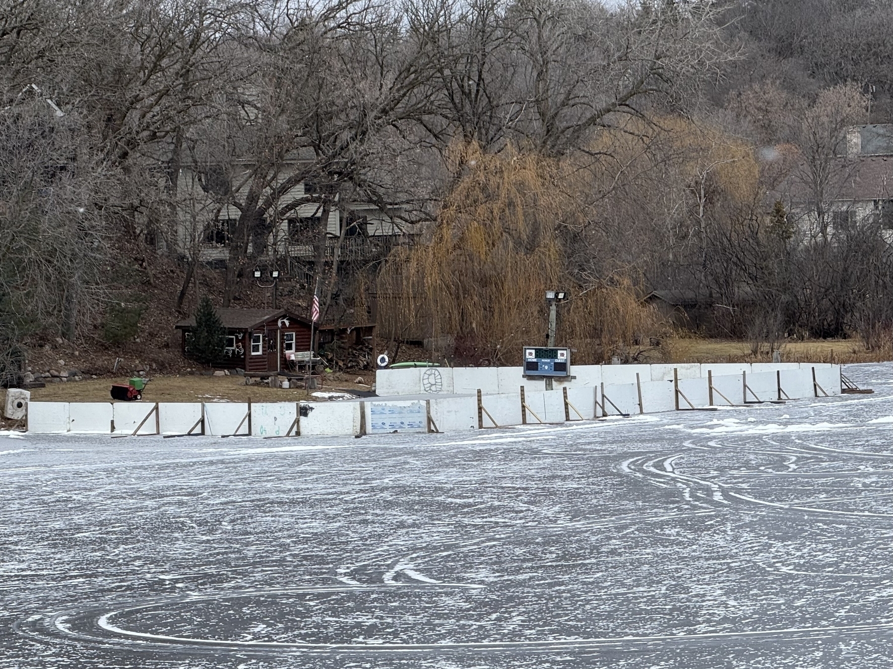 Ice rink surrounded by wooden boards sits frozen in a wintery wooded area. A small cabin with an American flag near rink’s edge. Snow-dusted trees and bare foliage fill the background.