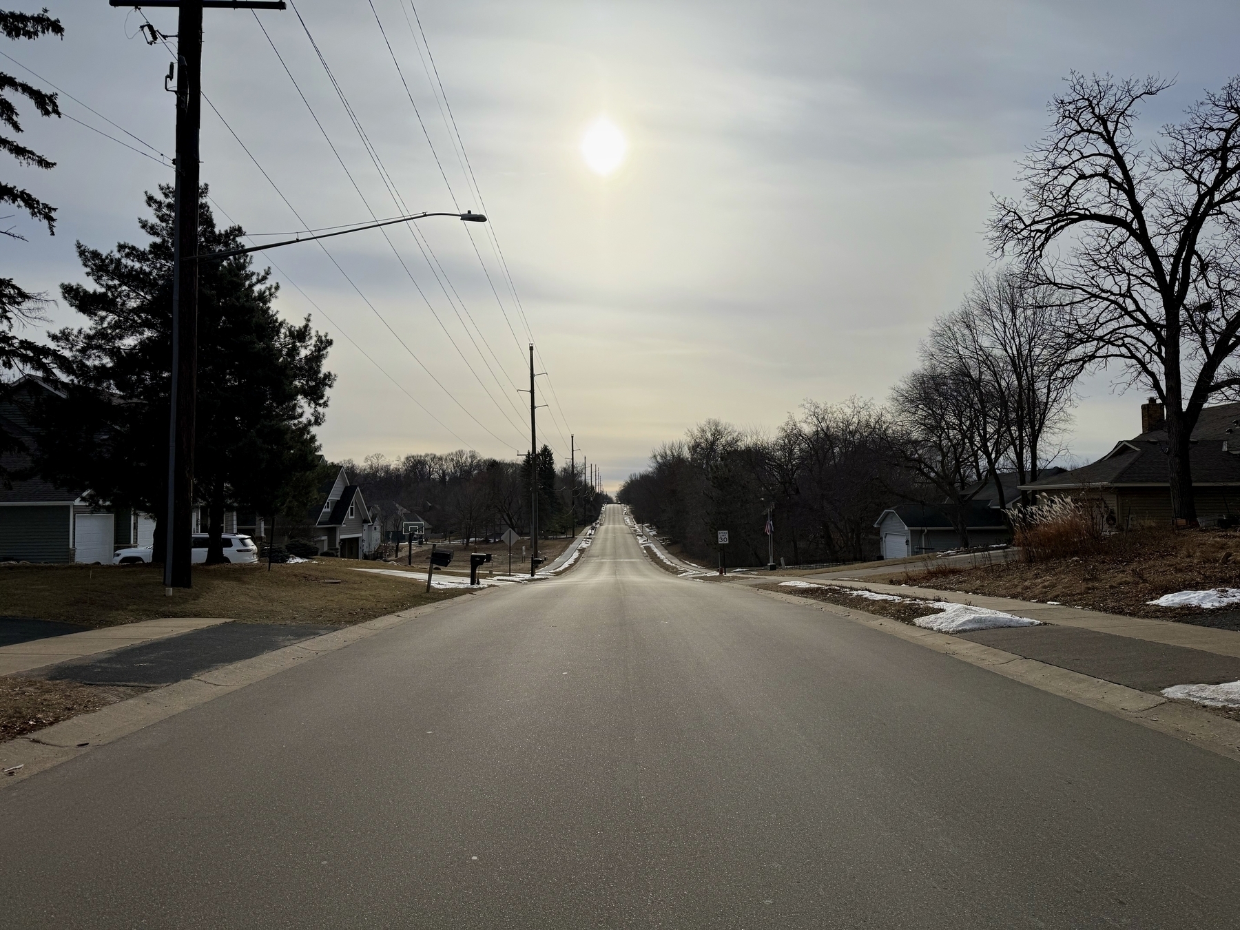 A long, empty road stretches into the distance under a cloudy sky, flanked by residential houses and leafless trees. The sun is visible, casting a soft light.