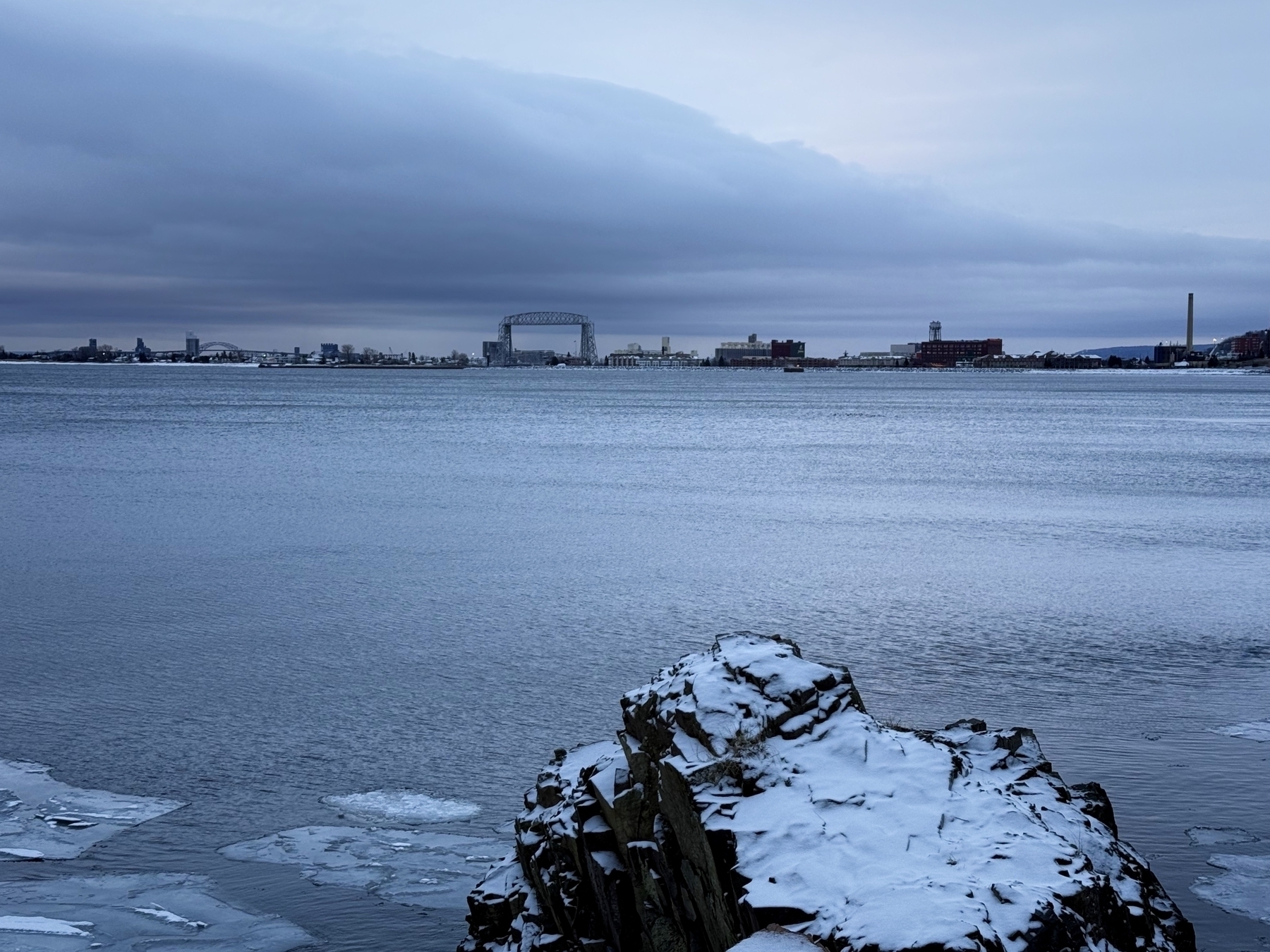 Snow-covered rocks rest at the edge of a calm, icy lake, with a large city bridge and skyline visible under a cloudy sky in the distance.