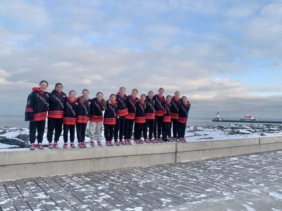 A group of thirteen children wearing matching black and red jackets stand closely on a snowy stone ledge near a rocky shoreline, with a sea and a distant lighthouse in the background.