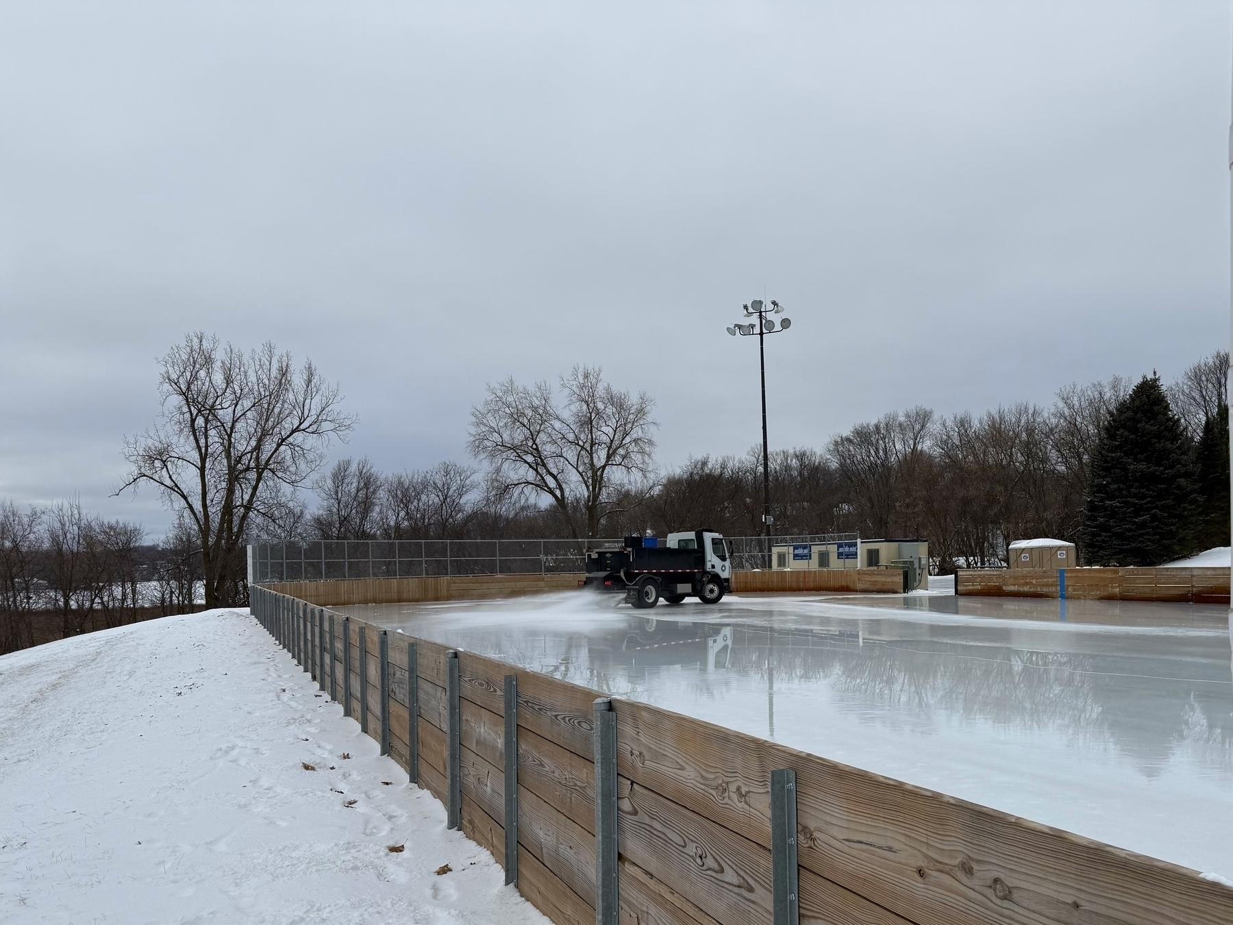 Truck smoothing ice on an outdoor rink surrounded by wooden barriers and snow, with leafless trees and a cloudy sky in the background.