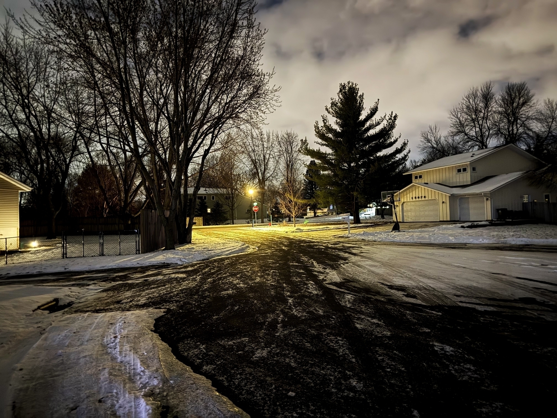 Street illuminated by lampposts casts a warm glow on icy road and snow-covered sidewalks, with bare trees and suburban houses in a quiet nighttime neighborhood.