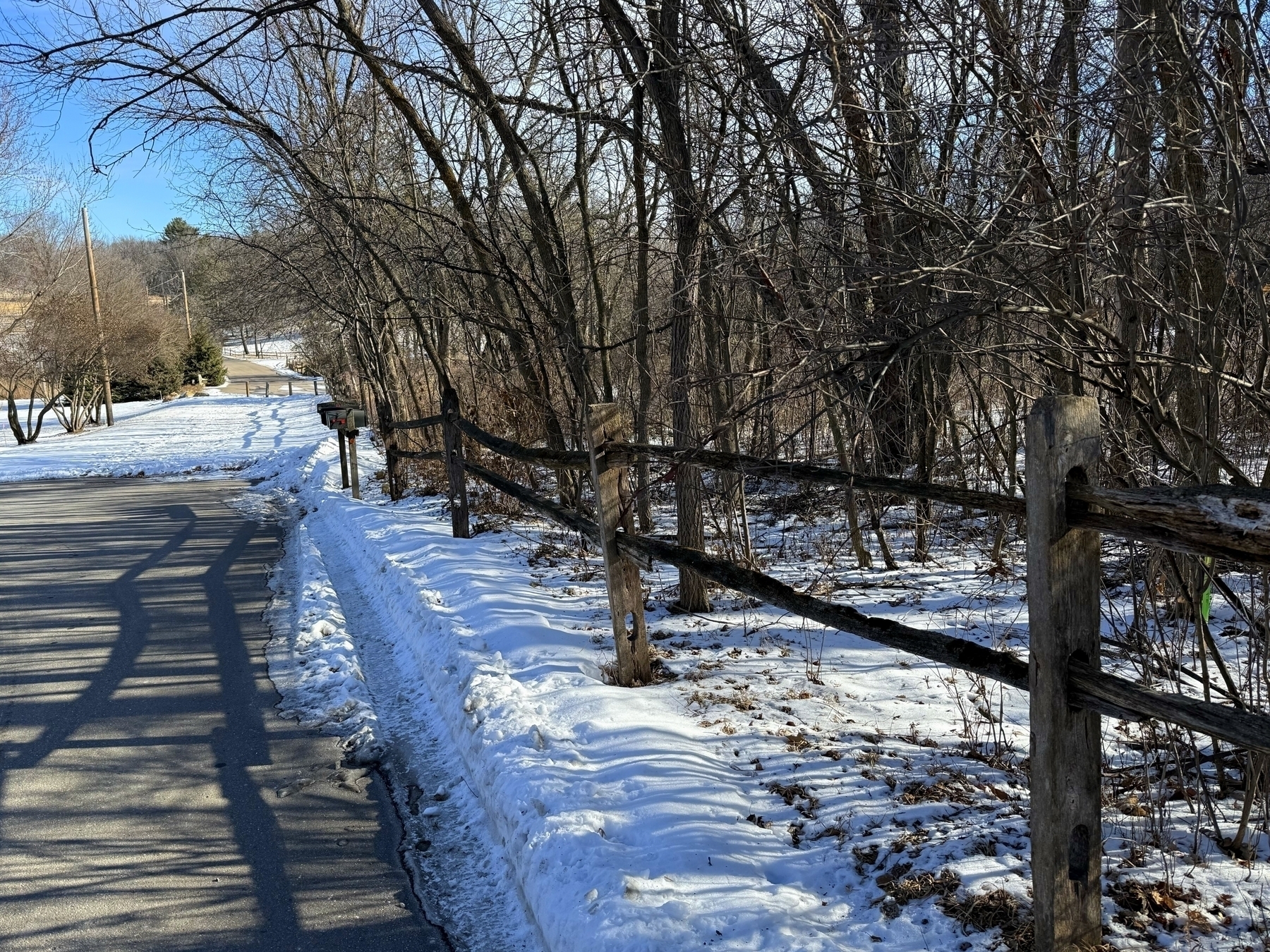 A rustic wooden fence runs parallel to a snowy road, bordered by bare trees, under clear blue skies in a rural winter landscape.