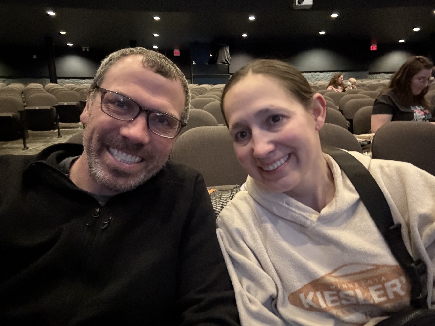 A smiling man and woman sit closely together in a theater with empty seats around them. The woman wears a hoodie with the text “Minnesota Kiesler.”