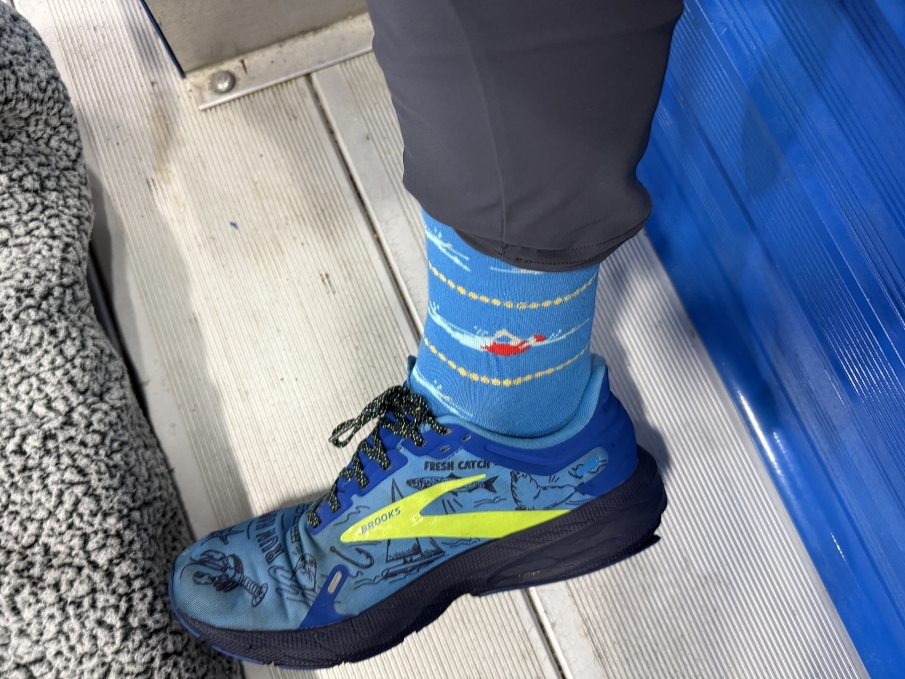 A blue shoe with lobster illustrations rests on a gray grooved surface. The wearer wears colorful socks depicting a swimming pool theme, standing beside a blue wall.