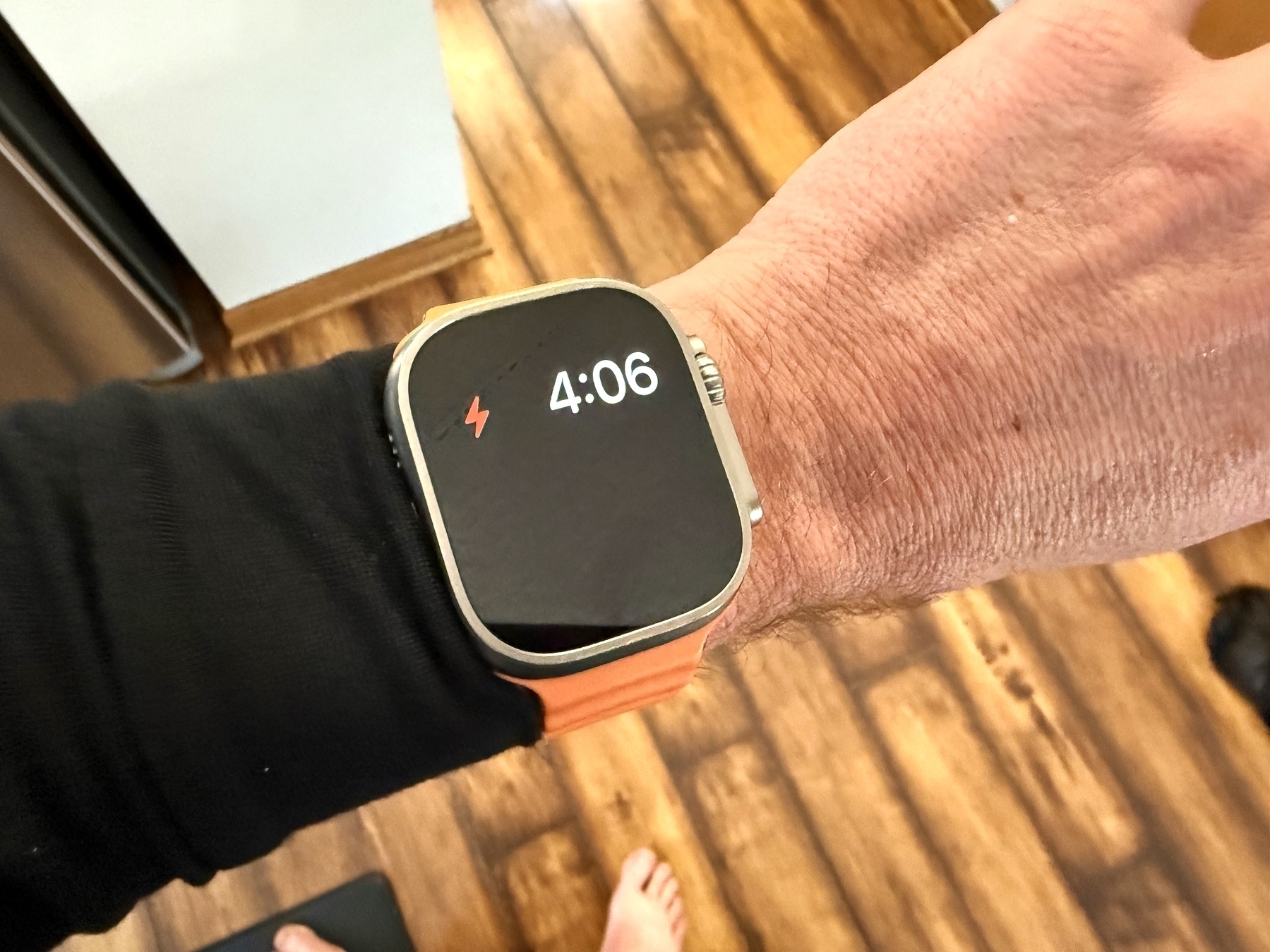 A smartwatch displays “4:06” with an orange band on a wrist. The wearer stands on wooden flooring, partially visible in a household interior.