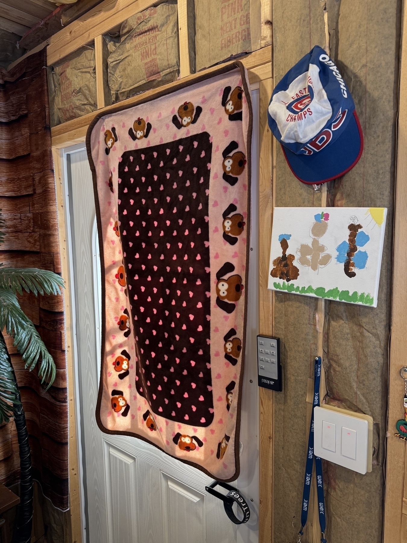 A blanket with cartoon bear designs and heart patterns hangs on a door. Nearby, a cap with “East Champs” is displayed, along with a child’s painting and light switches.