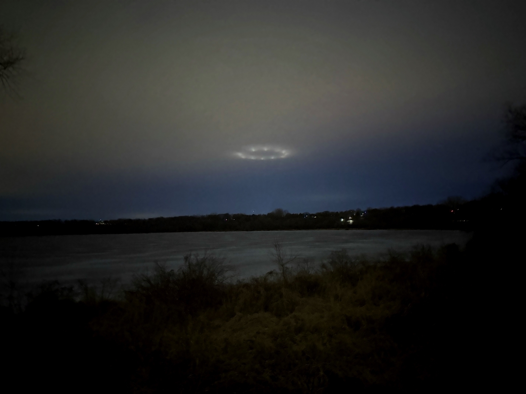 A nighttime landscape with a dark, cloudy sky. Below, there is a body of water surrounded by trees and vegetation along the shoreline. In the sky, there is a circular pattern of lights creating a glowing ring, illuminating the clouds around it. The overall atmosphere is mysterious and serene.