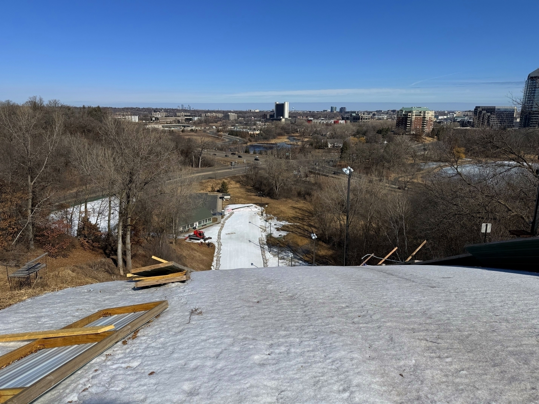 A view from the top of a snow-covered hill looking down a ski slope. The slope is surrounded by bare trees, with patches of snow visible on the hillside. A building and a red snow grooming machine are located at the base of the slope. In the background, there’s a cityscape that includes several buildings and clear blue sky. The scene is bright and sunny, indicating a clear day.