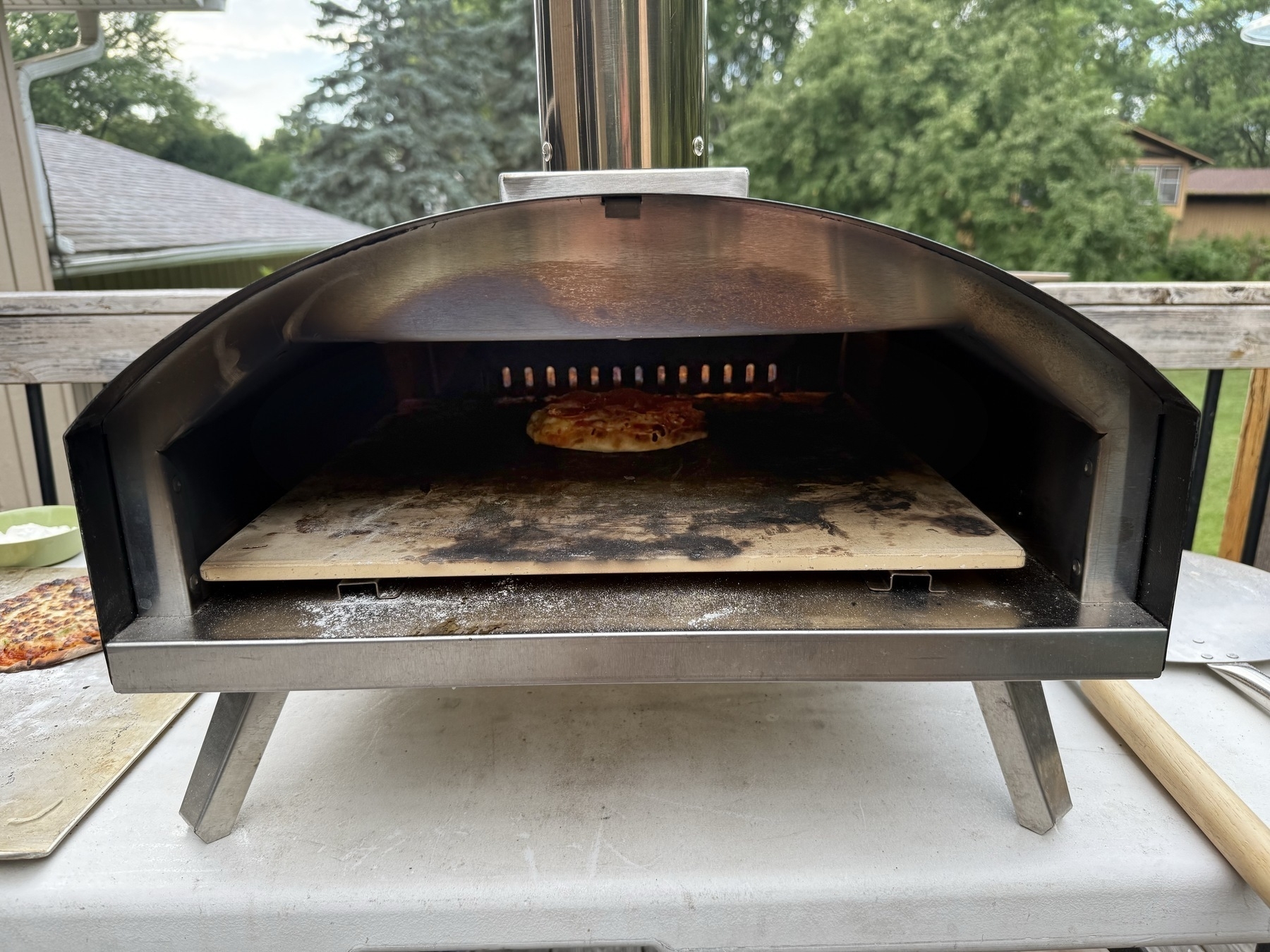 A wood-fired pizza oven with a partially cooked pizza inside, set outdoors on a table. Surrounding greenery and a house are visible in the background.