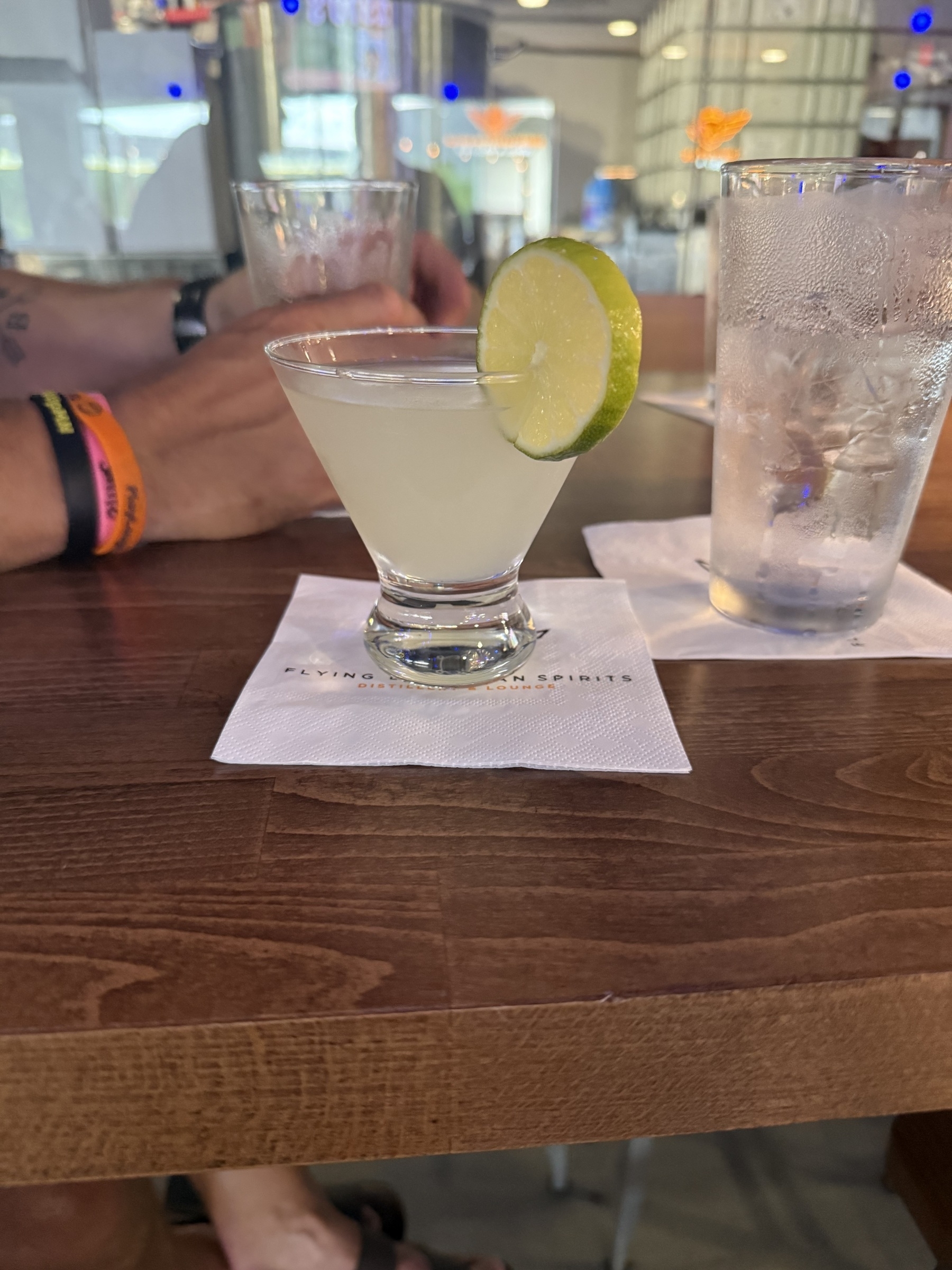 A cocktail garnished with a lime slice sits on a napkin on a wooden table, surrounded by other drinks and glasses in a bar setting.