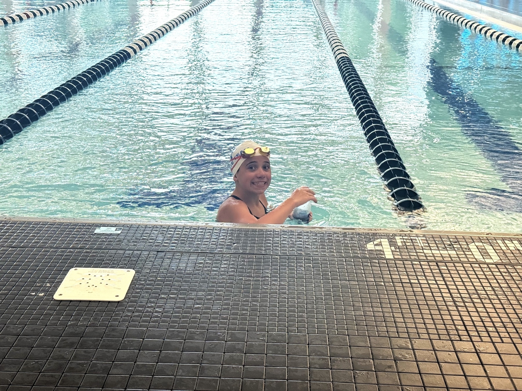 A swimmer in a black swimsuit and swim cap smiles while resting at the edge of an indoor pool, holding a water bottle. Lane dividers stretch across the pool in the background.