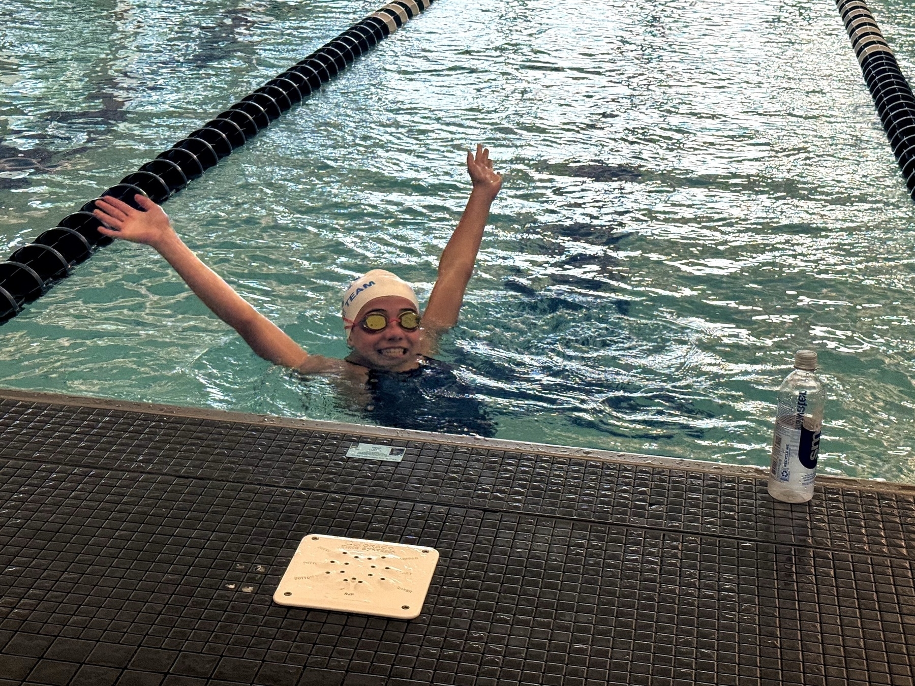A swimmer with a cap and goggles joyfully raises their arms in a pool, near a bottle of water on the deck.