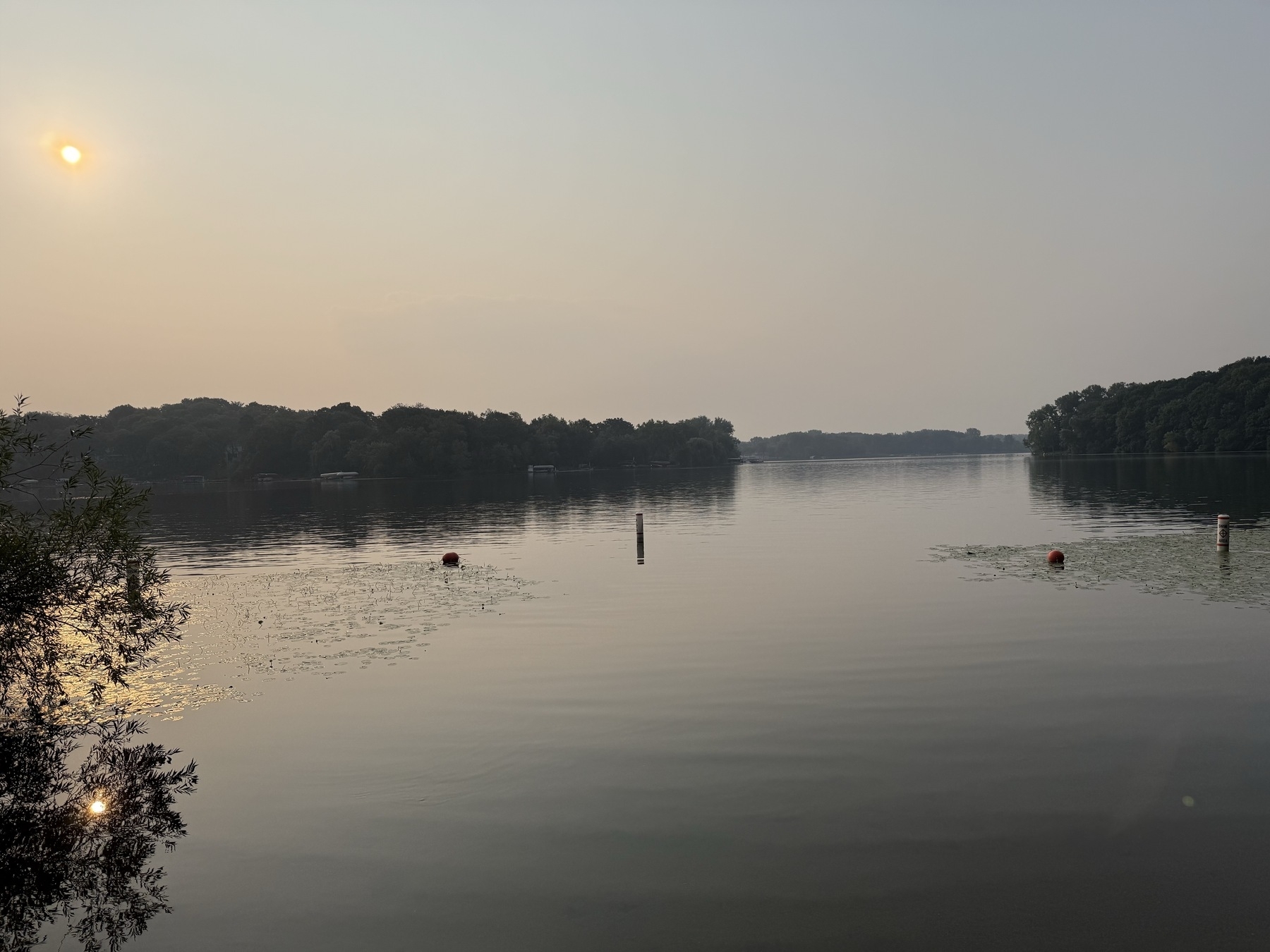 Tranquil lake with a hazy sky during sunrise, surrounded by dense trees. The water reflects the soft sunlight, and a few buoys are visible in the distance.
