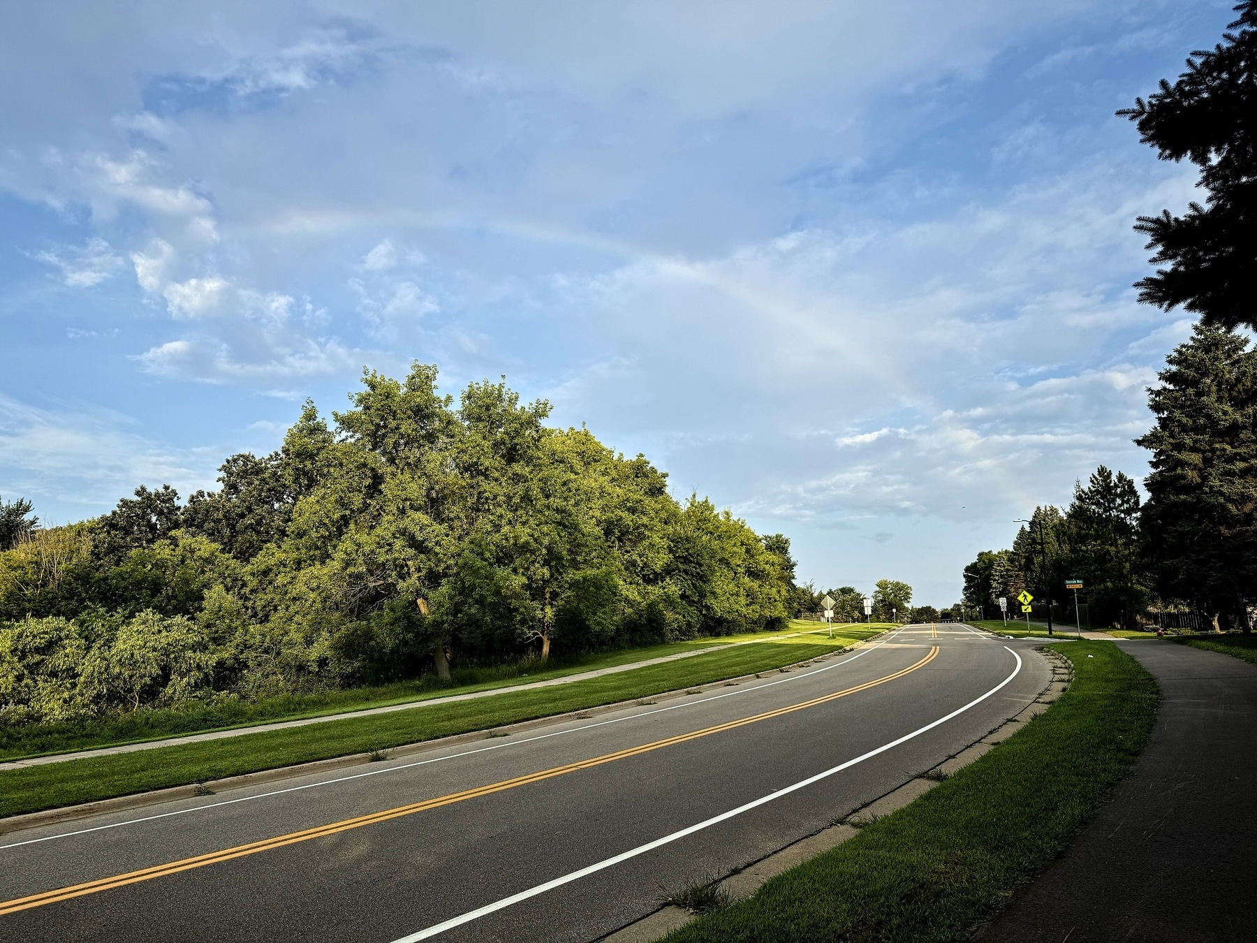A curved road bordered by lush green trees under a blue sky with scattered clouds and a faint rainbow.